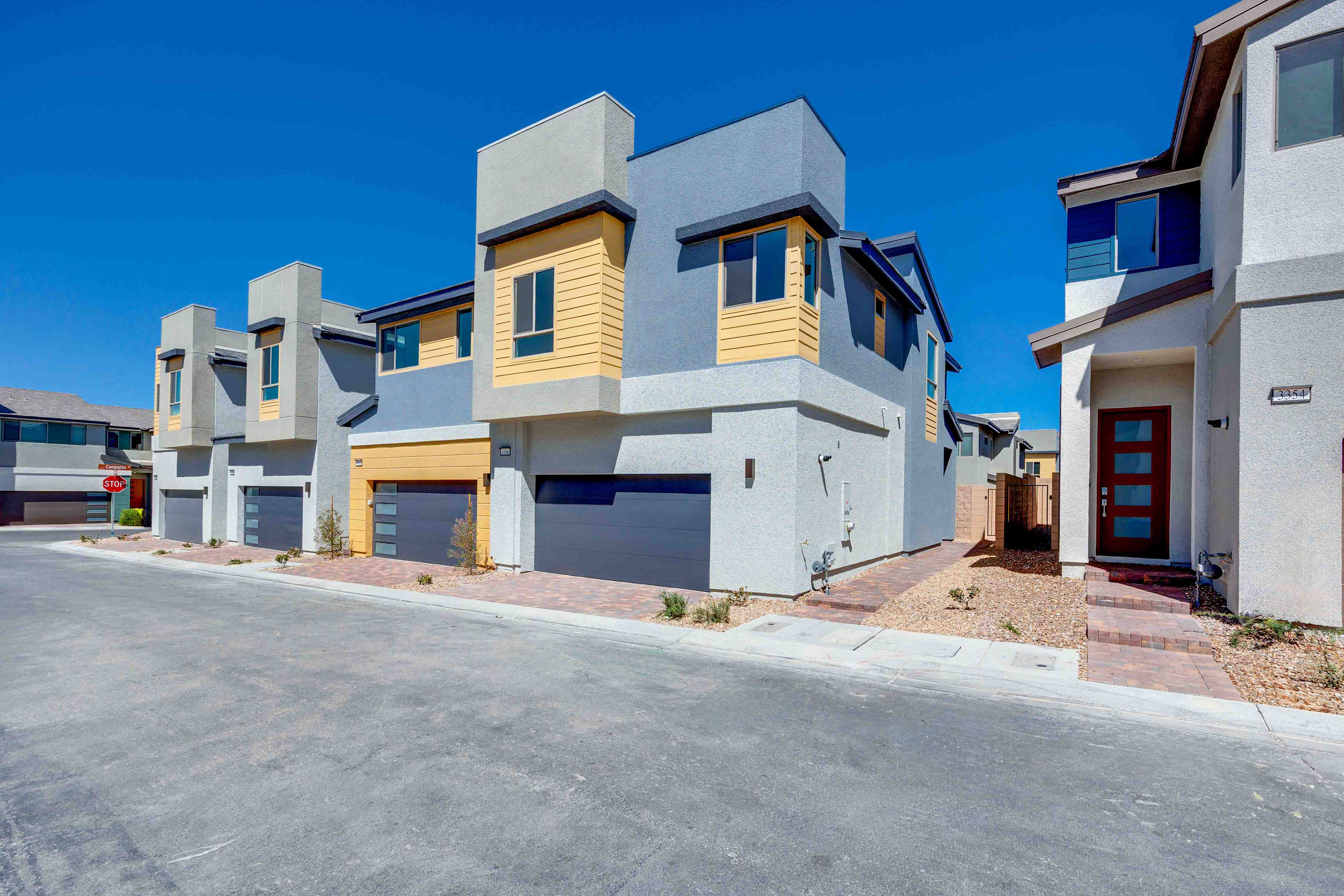 Modern townhouses with gray garages and colorful accents line a paved street under a clear blue sky.