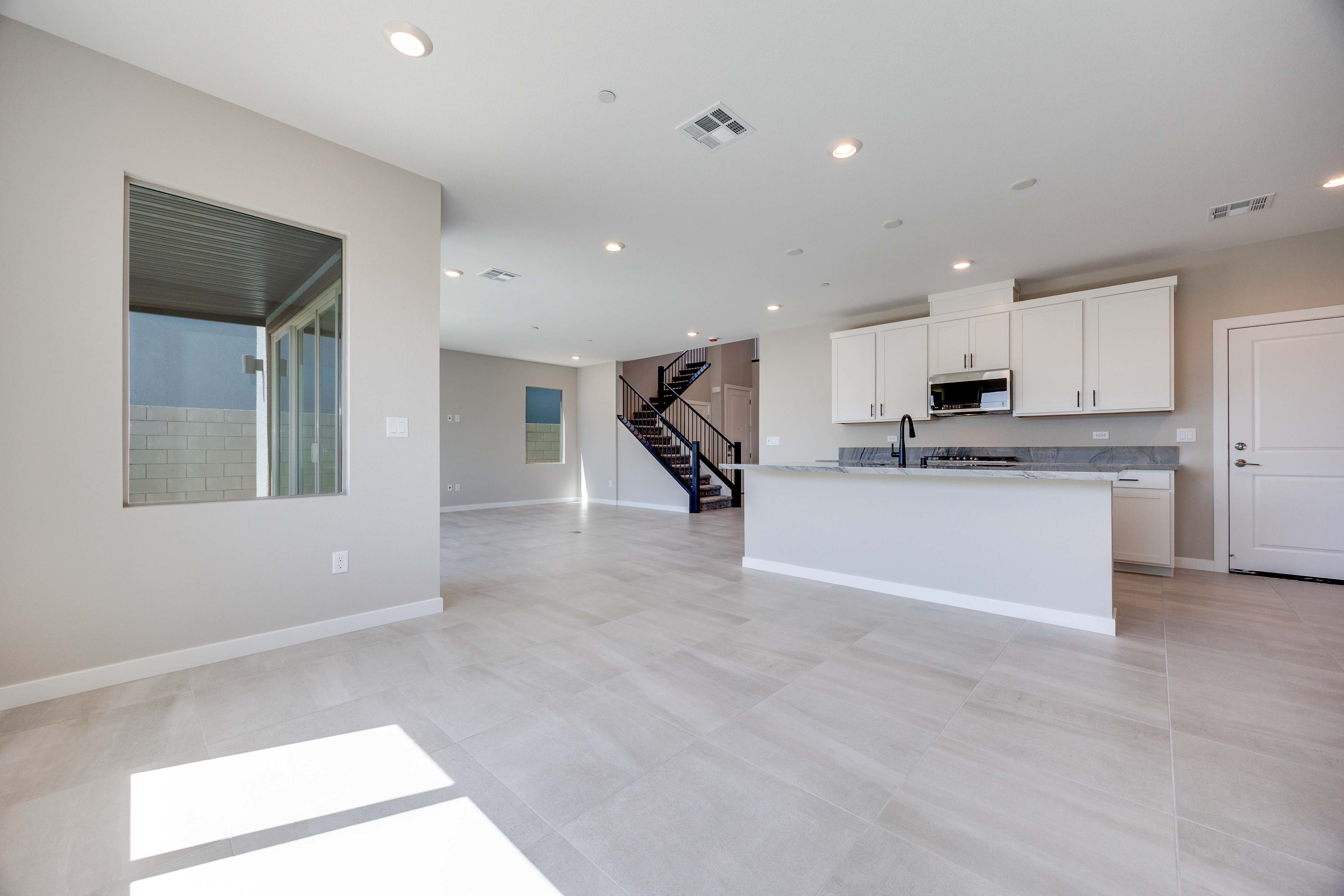 Sunlight streams across the light-tiled floor of an open-concept living area with a modern kitchen and a staircase in the background.