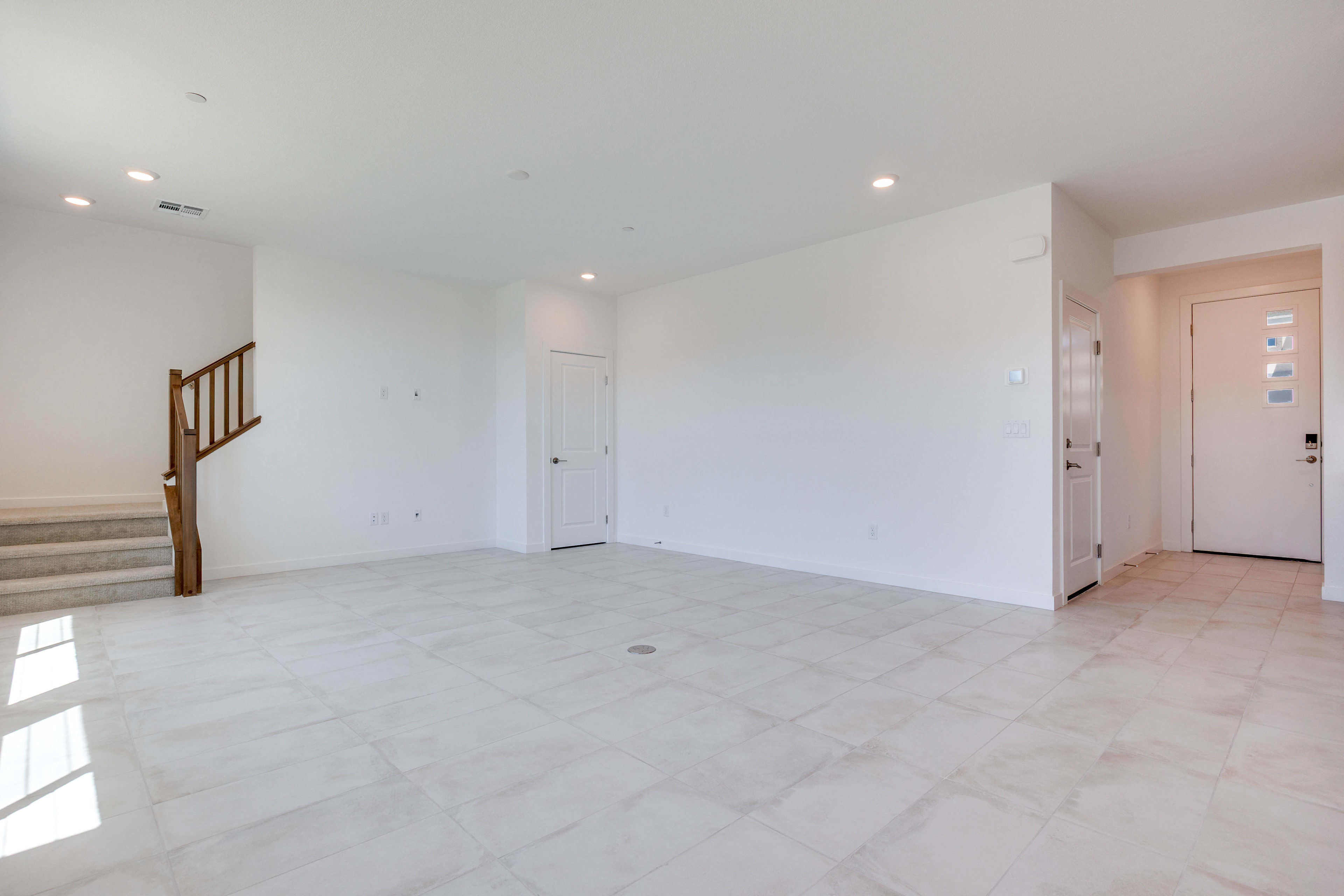 A bright, empty living room with white tiled floors in the foreground and two doors and a staircase in the background.