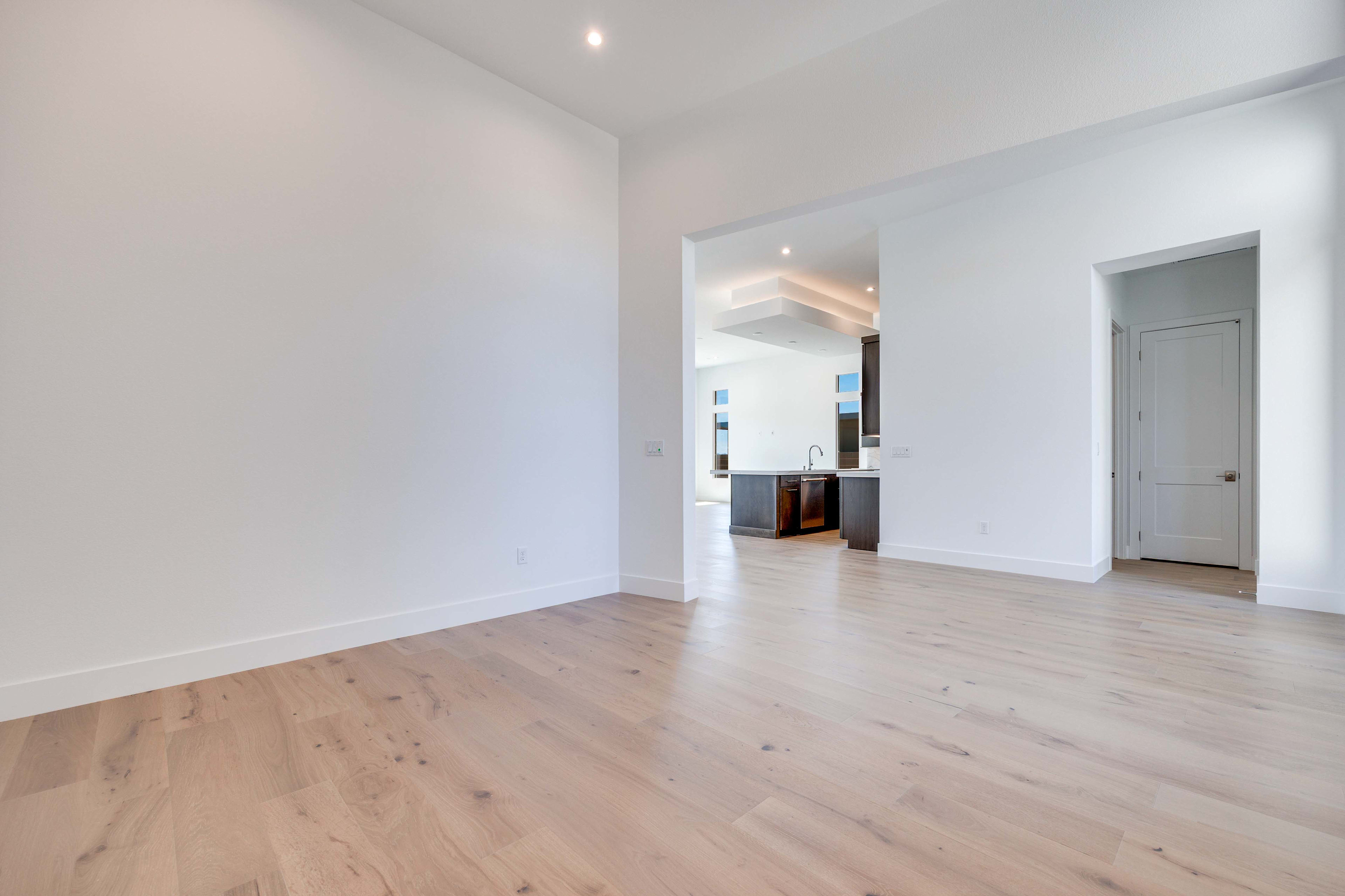 A bright, empty room with light wood floors in the foreground opens to a modern kitchen with dark cabinets and a sleek island in the background.