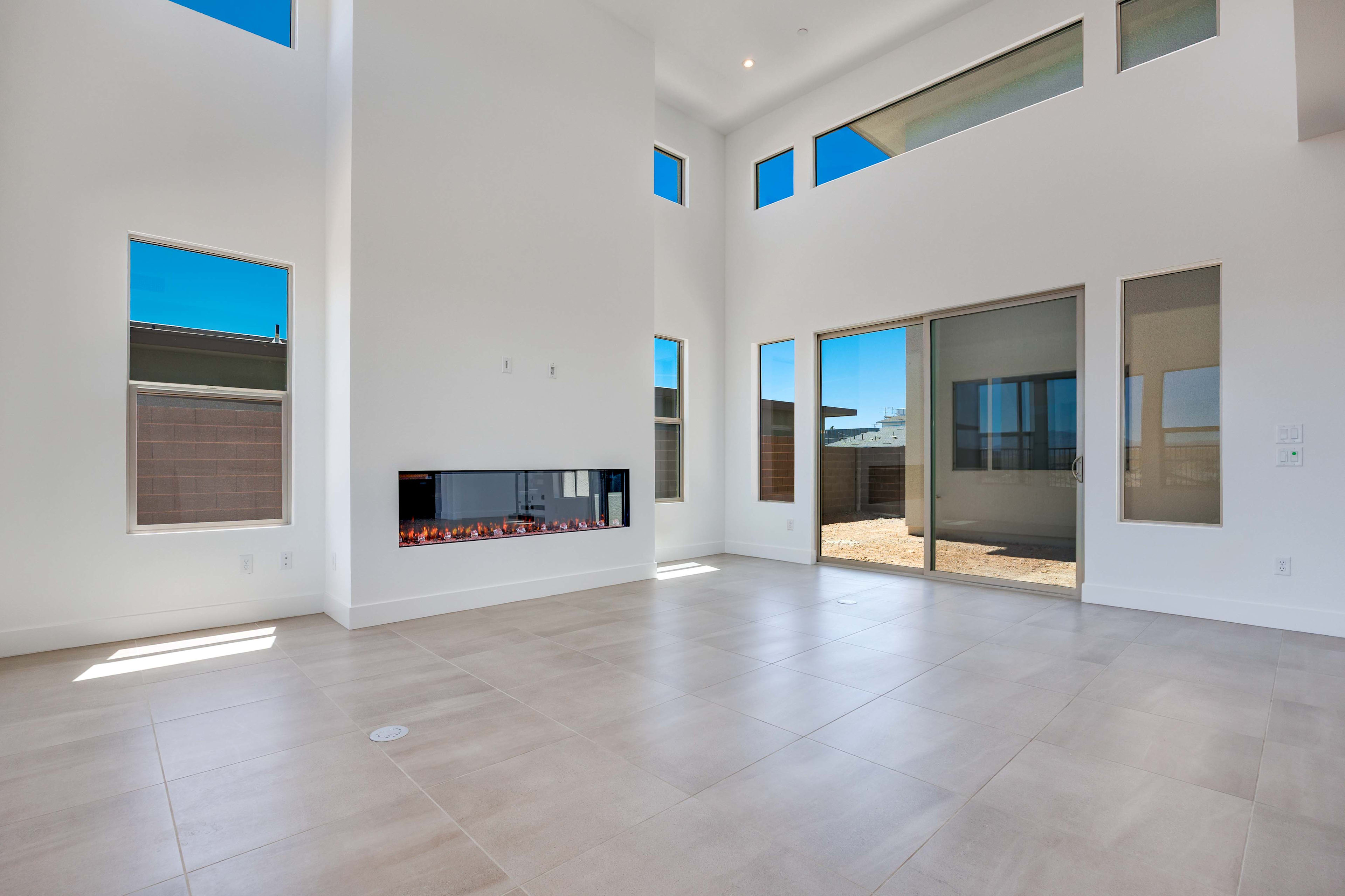 A spacious, modern living room with a sleek fireplace in the foreground opens to a sunny outdoor patio in the background.