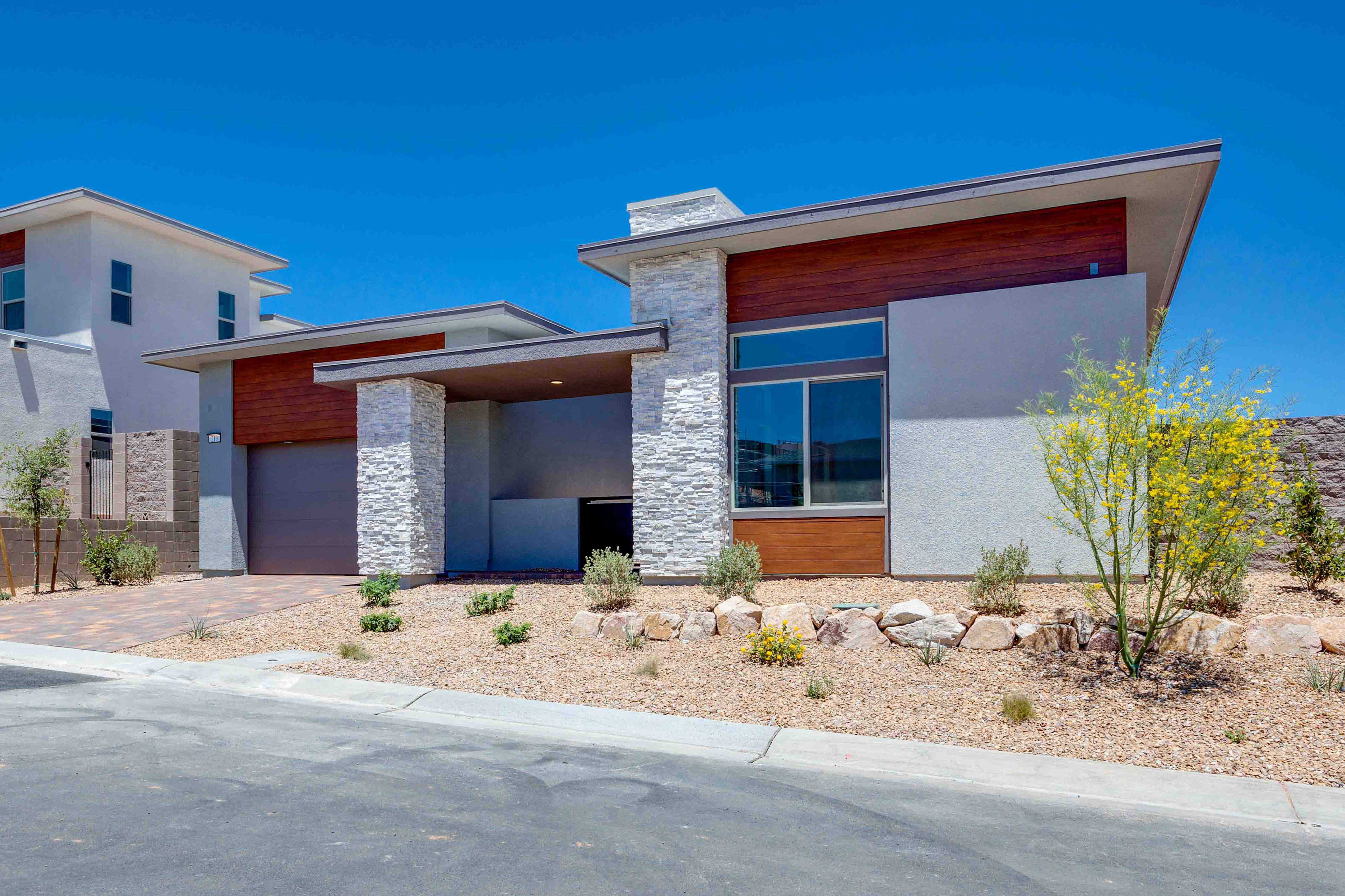 A modern, multi-level house with stone and wood accents sits behind a gravel yard with sparse landscaping, under a clear blue sky.