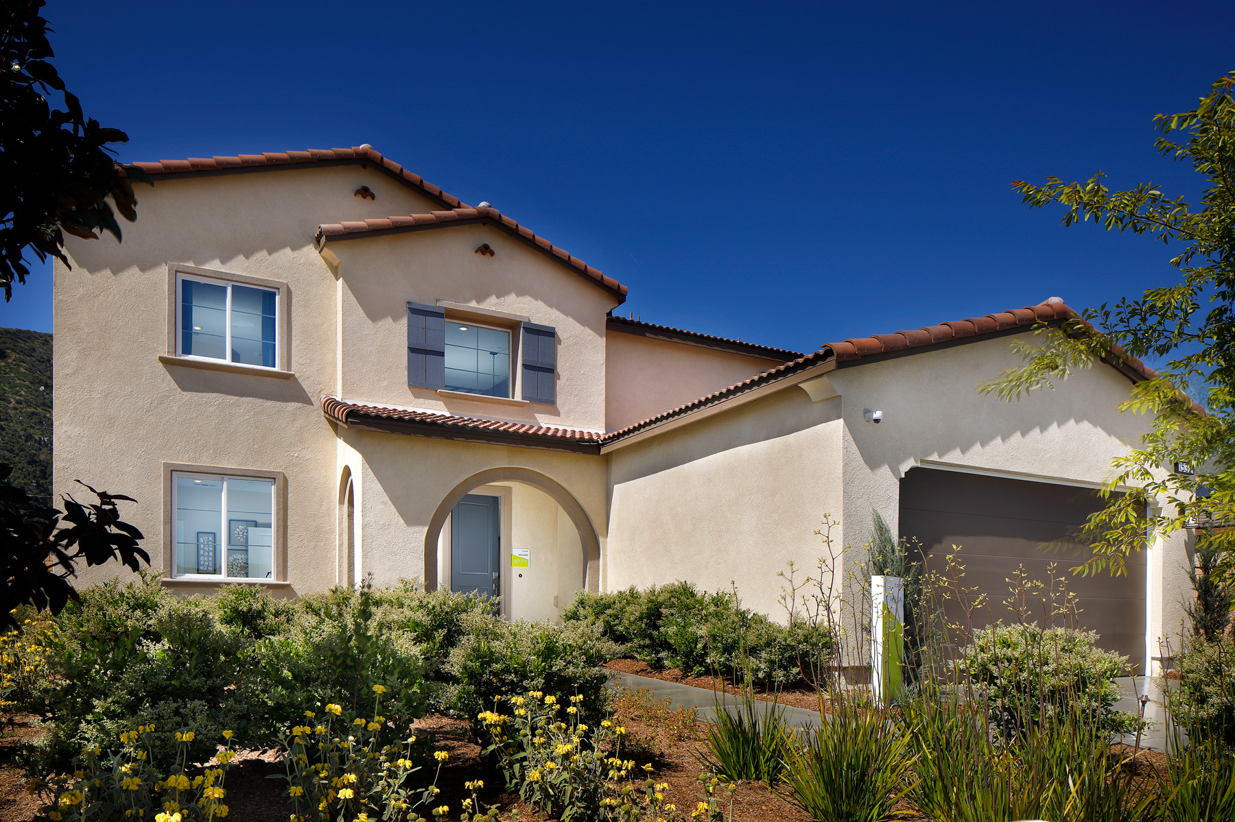 A two-story Mediterranean-style house with a tiled roof, surrounded by lush greenery and a well-manicured garden in the foreground, set against a clear blue sky.