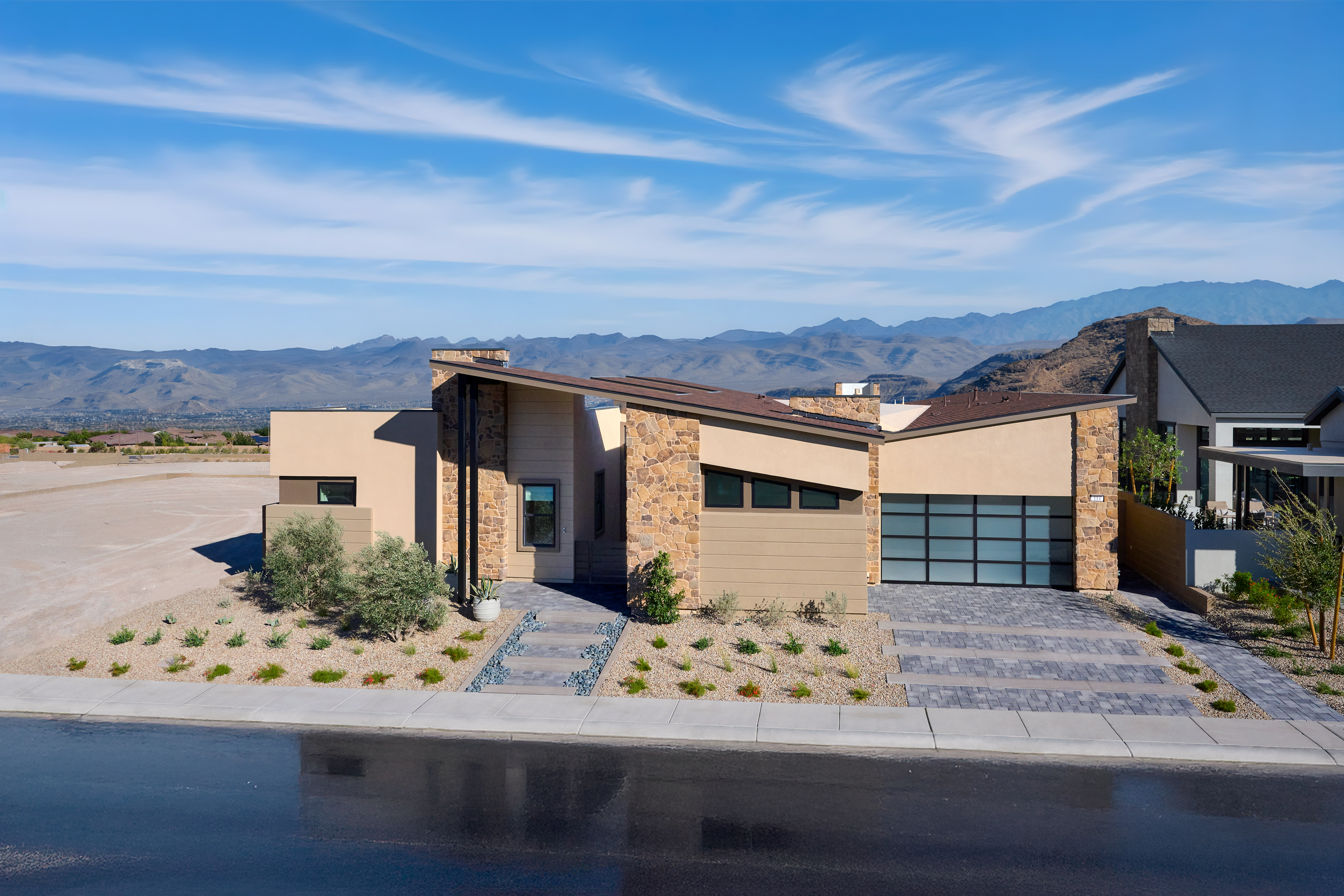 A modern, two-story house with a stone and stucco exterior, surrounded by a paved driveway and landscaped yard, set against a backdrop of mountains and a blue sky with wispy clouds.