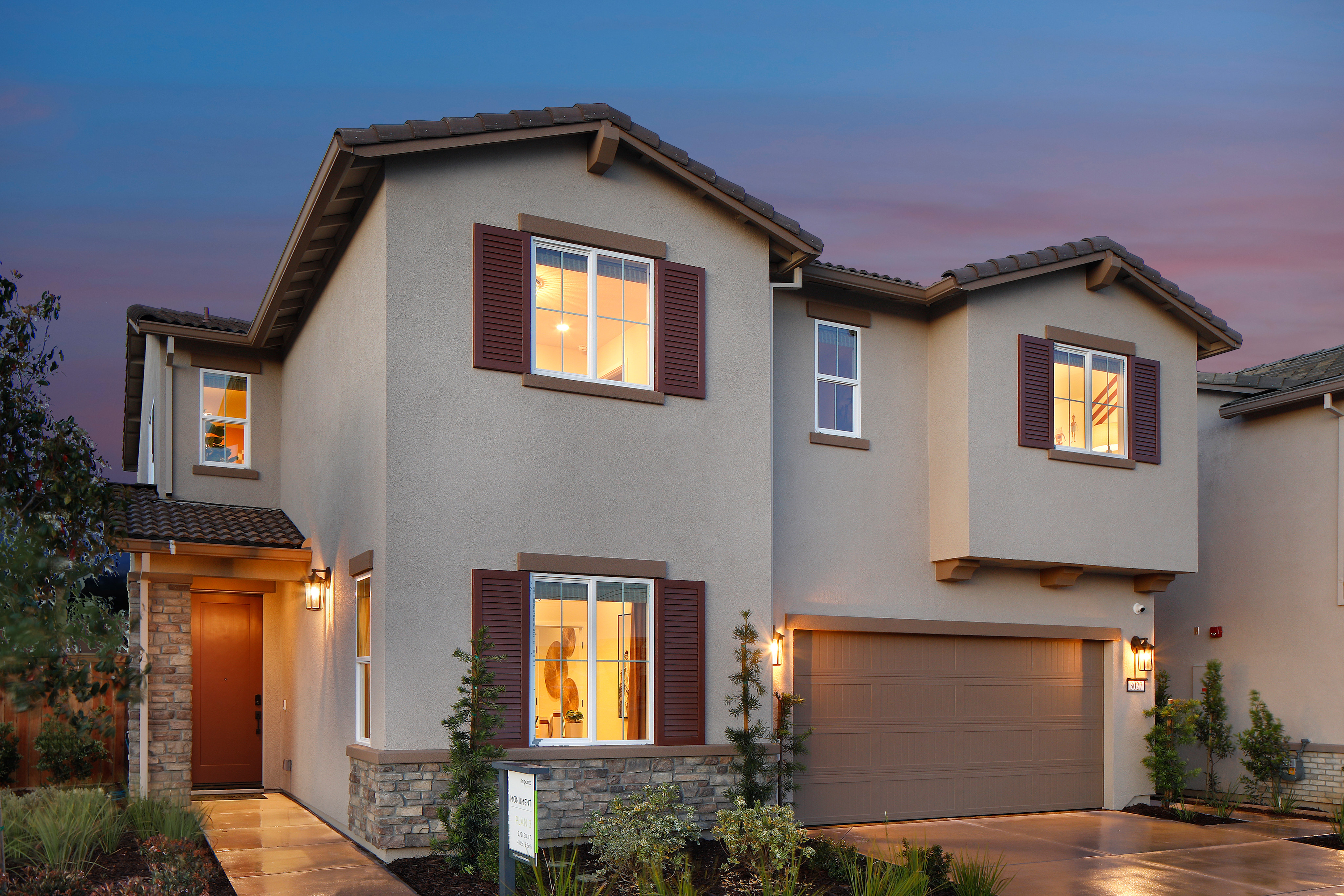 A two-story residential house with a garage, surrounded by landscaping and illuminated at dusk against a colorful sky.
