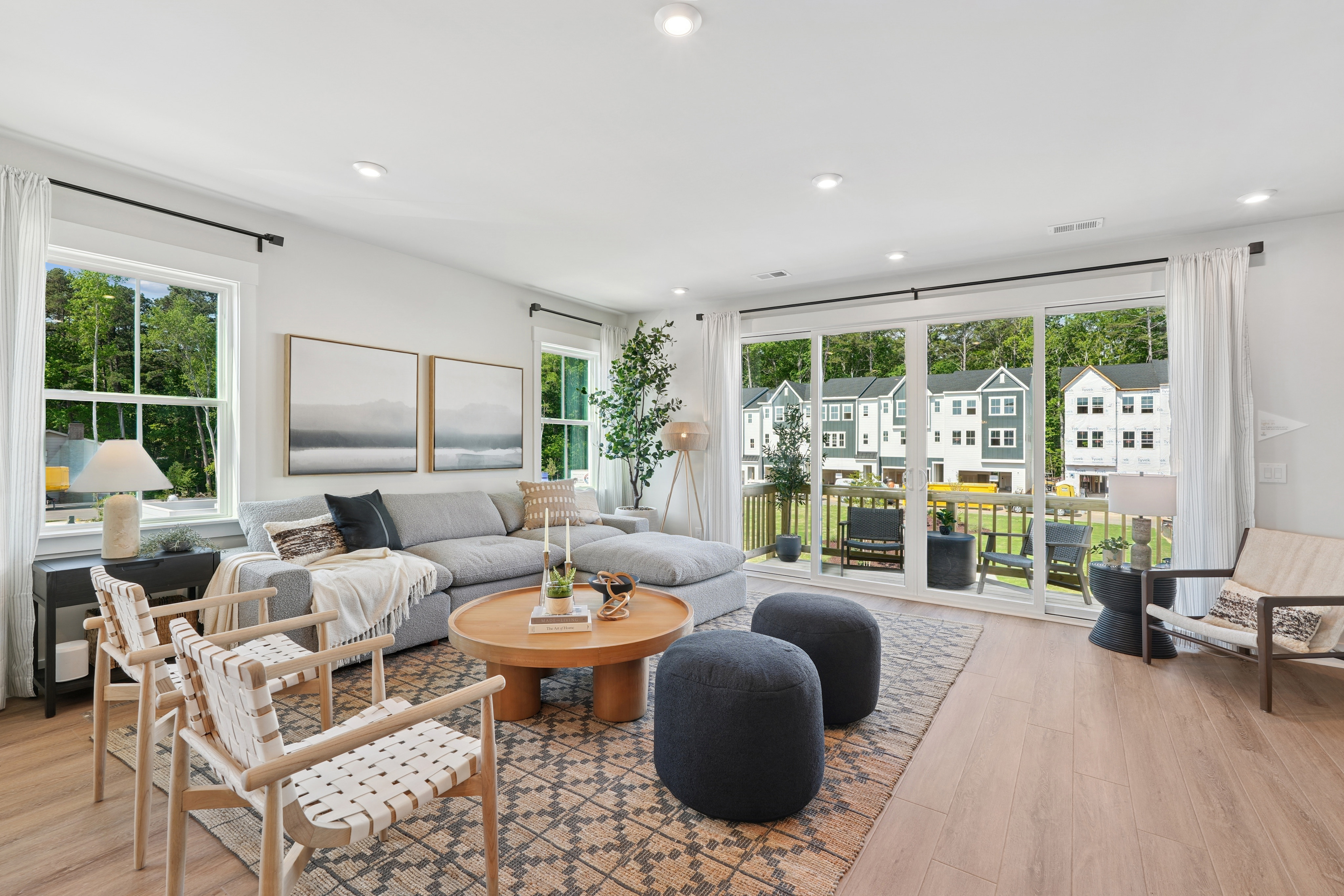 A cozy and modern living room with large windows overlooking a residential neighborhood, featuring a gray sectional sofa, a round wooden coffee table, and various decorative elements.