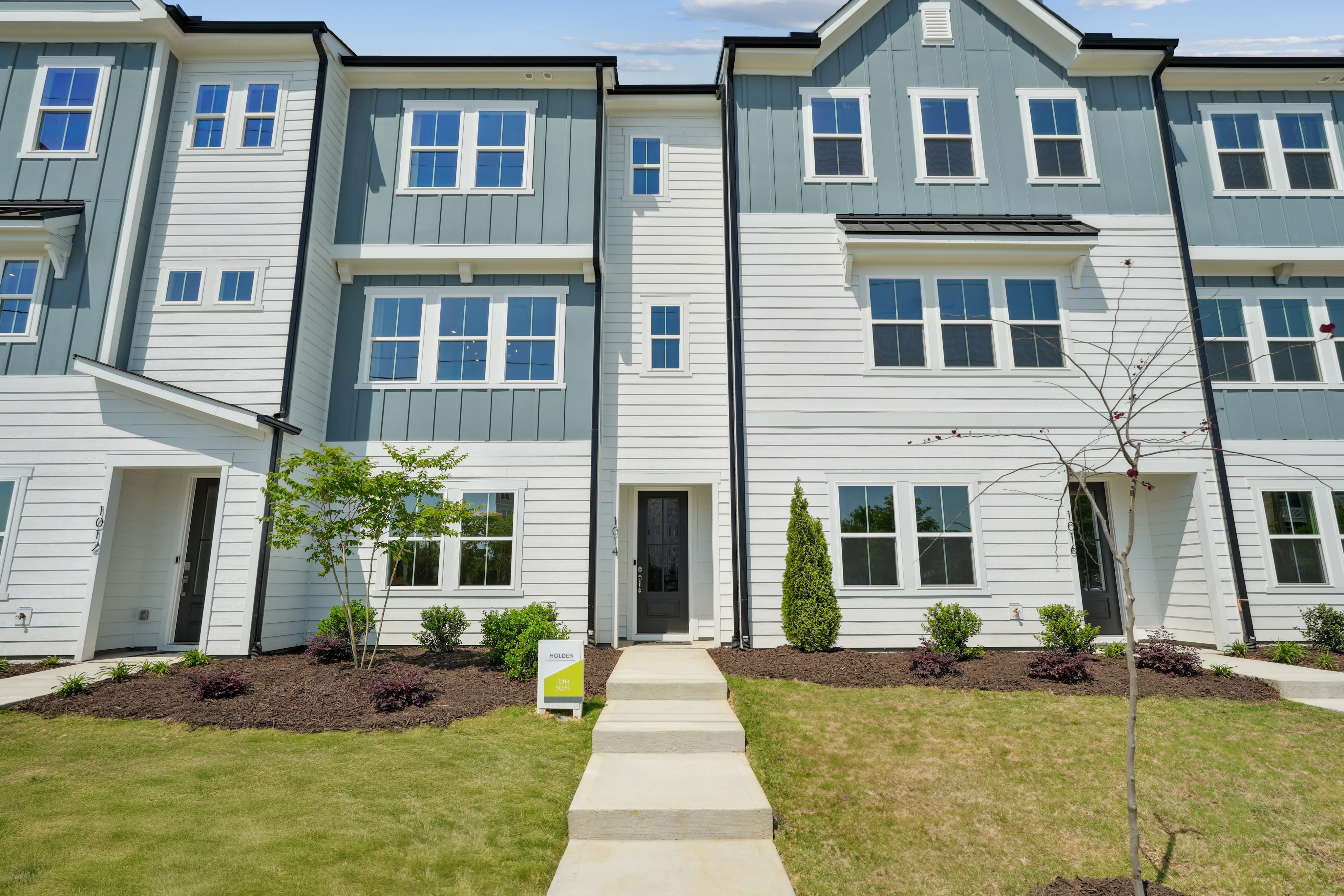 A multi-story townhouse community with a well-manicured lawn and landscaping in the foreground, featuring a mix of siding colors and architectural styles.