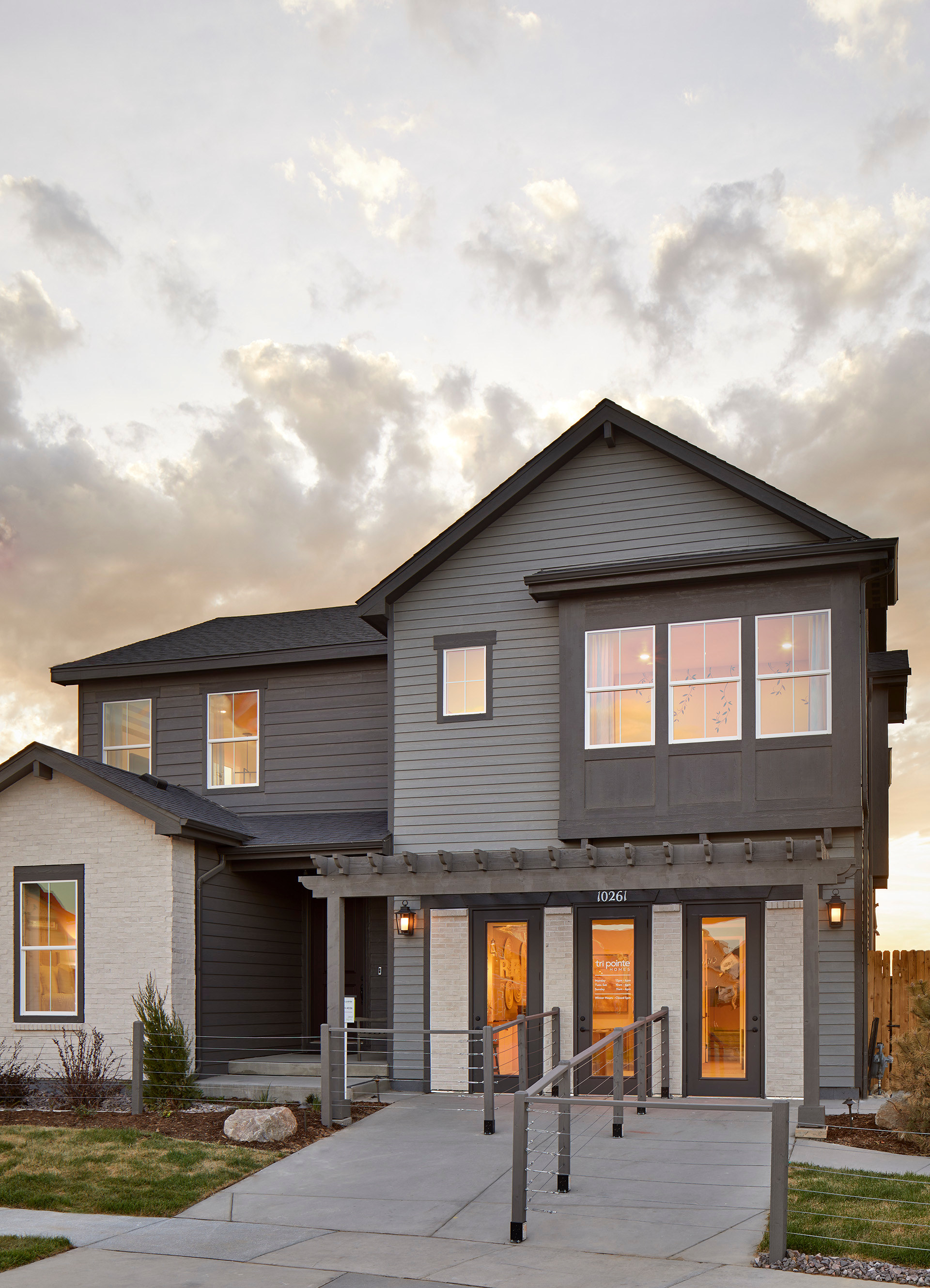 A two-story residential building with a porch and large windows, set against a cloudy sky with a grassy foreground.