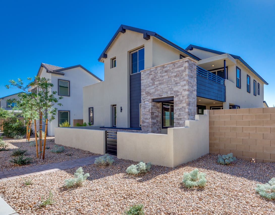 A modern, two-story residential building with a stone facade, surrounded by a gravel yard and desert landscaping against a clear blue sky.