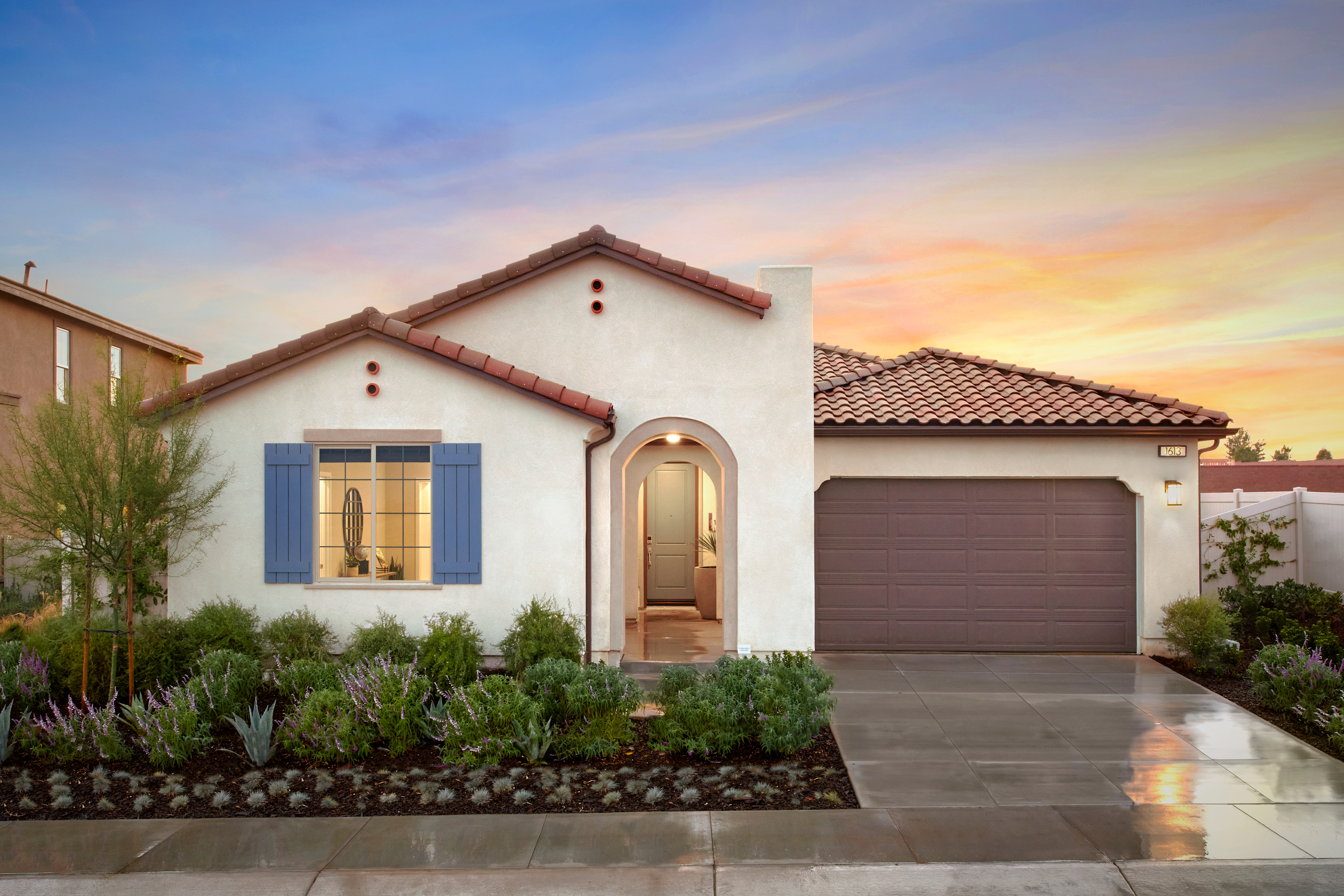A single-story Mediterranean-style house with a tiled roof, surrounded by lush landscaping, set against a vibrant sunset sky.