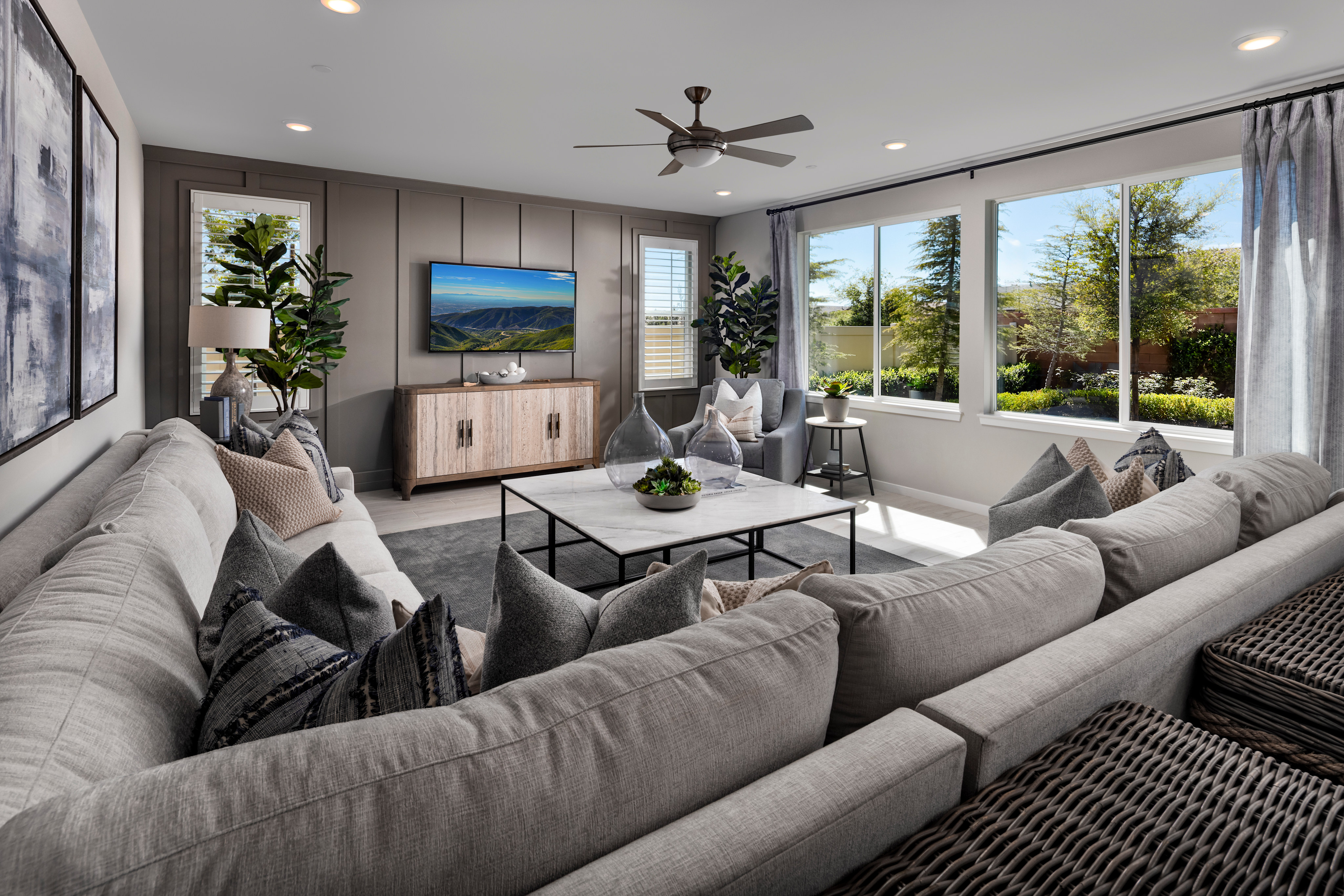 A cozy and modern living room with plush gray sofas, a large TV, and a view of the lush outdoor scenery through the windows.