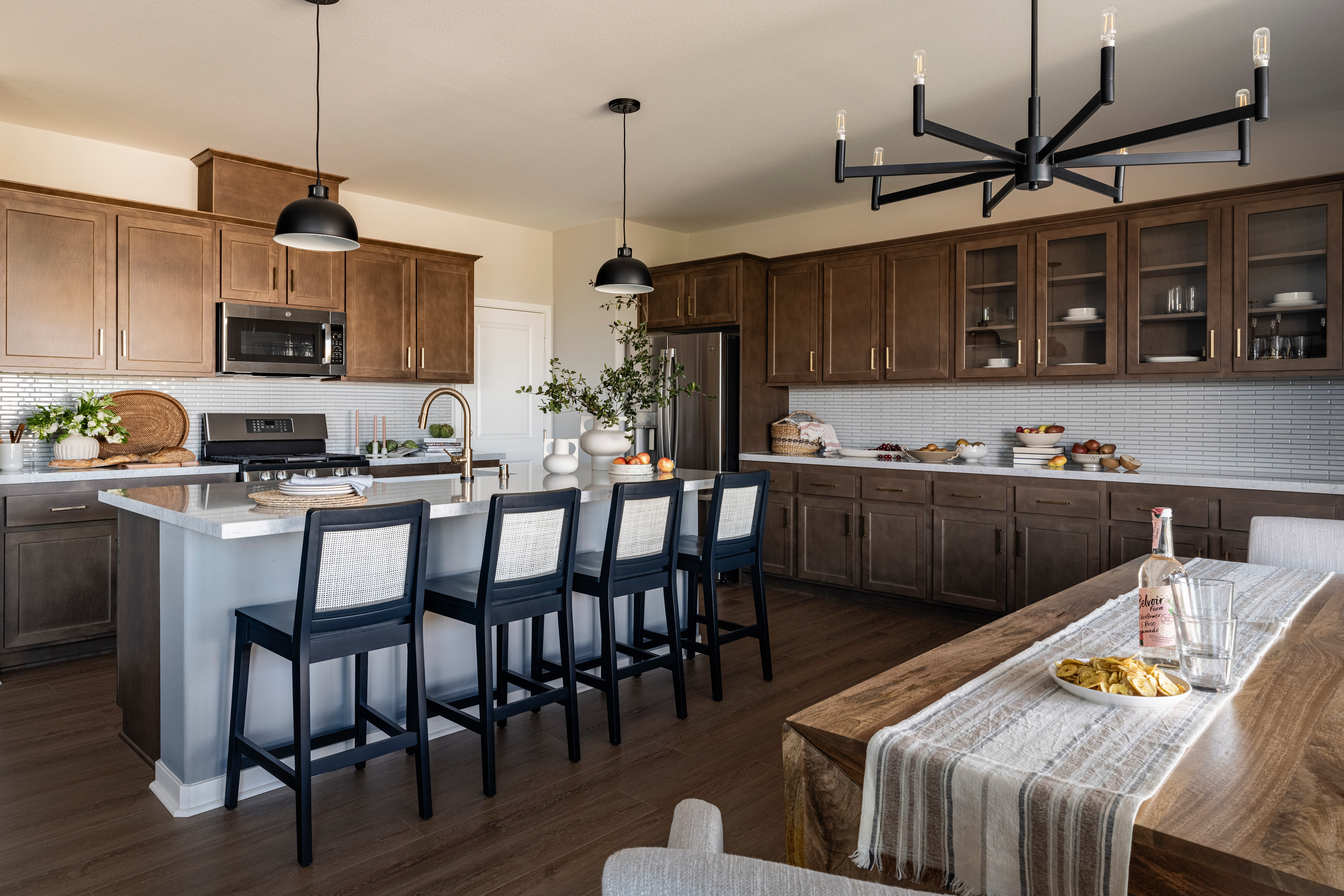 A modern and spacious kitchen with wooden cabinets, a large island with bar stools, and a chandelier overhead, set against a backdrop of a dining area with a table and chairs.