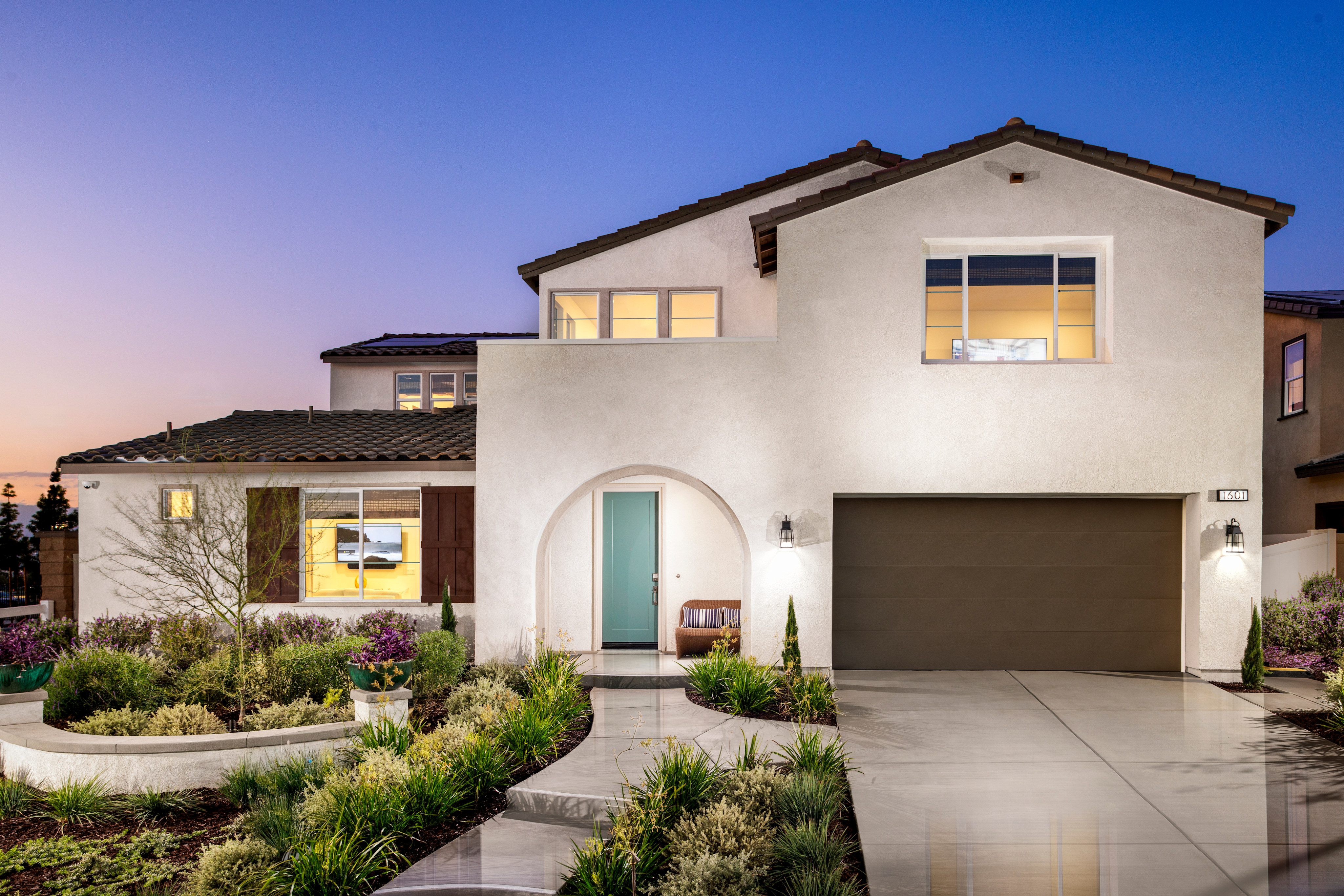 A modern, two-story white stucco house with a tiled roof, surrounded by a well-landscaped yard featuring a paved driveway, steps, and various plants and shrubs, set against a clear blue sky at sunset.