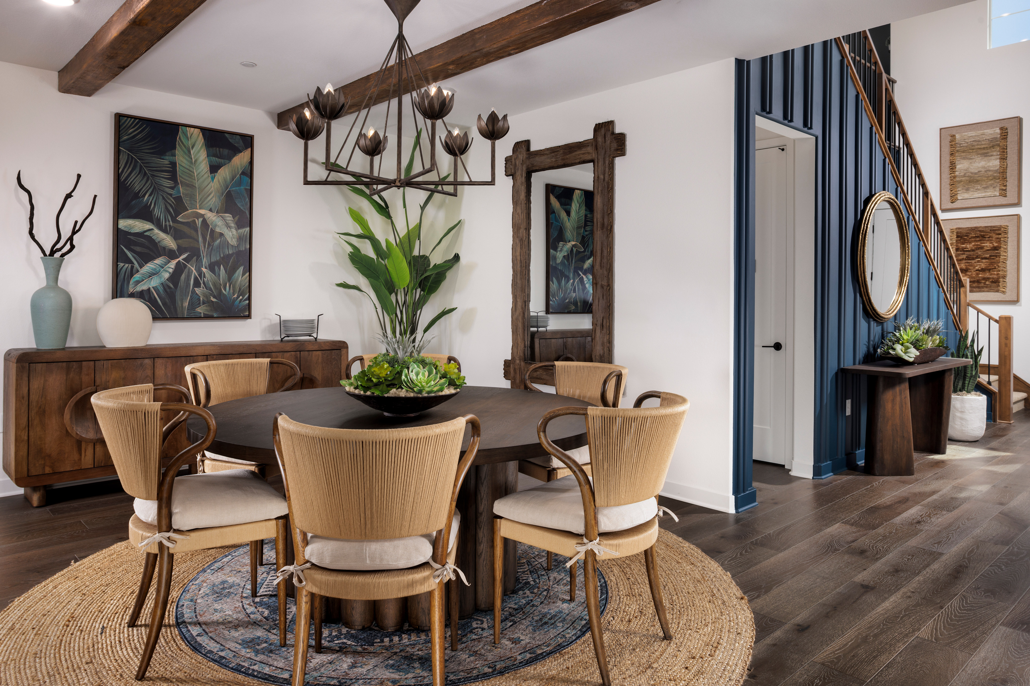 A cozy and stylish dining room with a wooden table surrounded by beige chairs, featuring a large tropical plant artwork, wooden beams, and a mirror on the wall.
