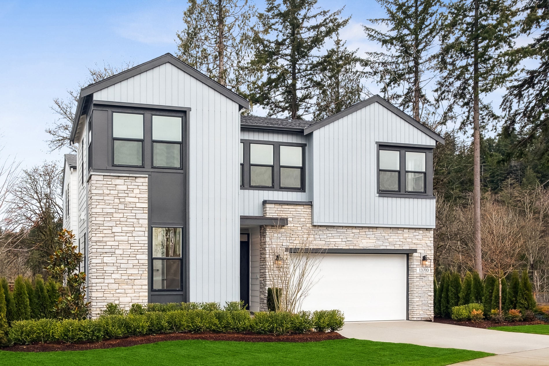 A modern two-story house with a white exterior, stone accents, and a garage door stands amidst a well-landscaped yard with lush greenery and trees in the background.