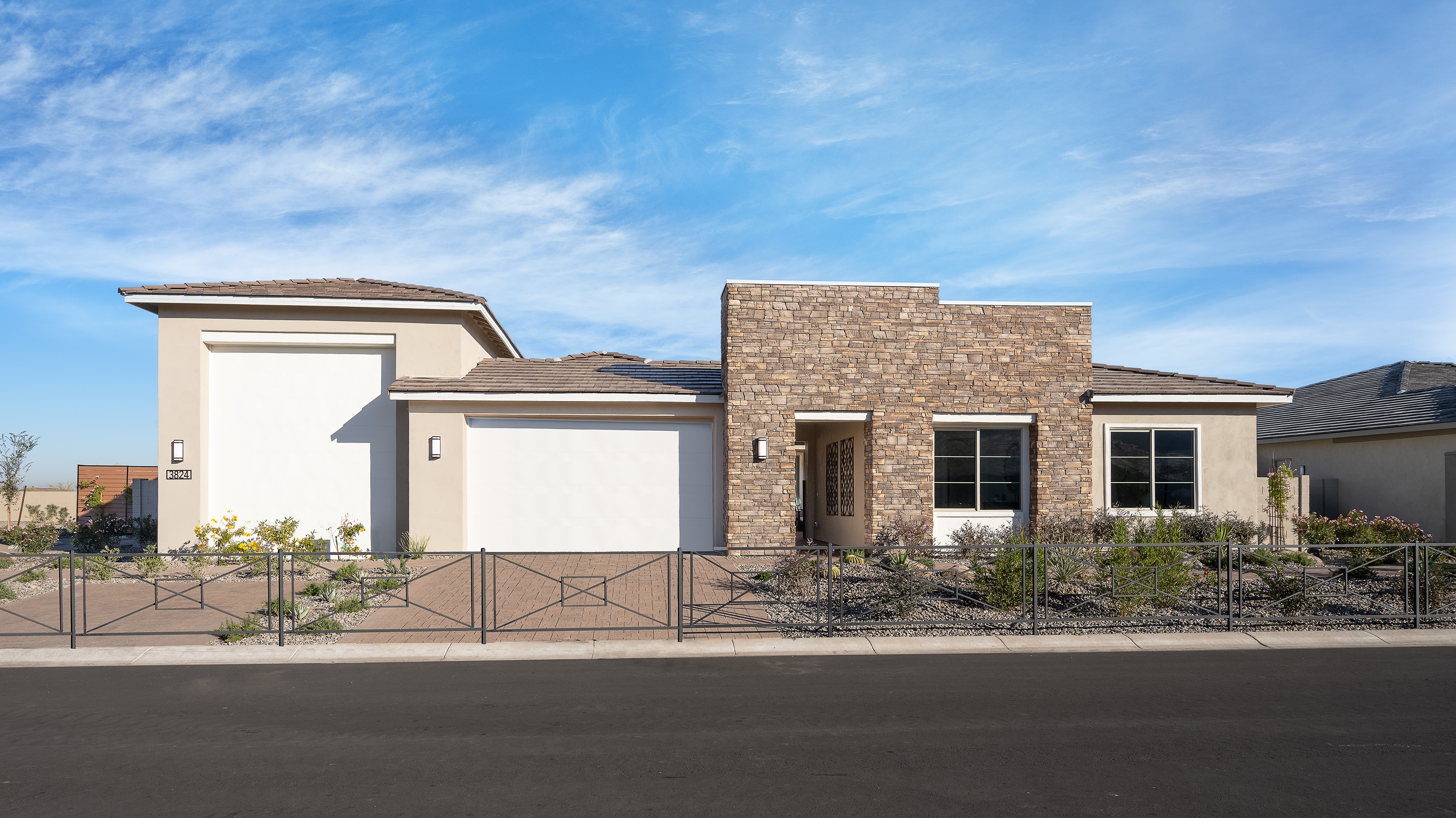 A modern, single-story house with a stone facade, a tiled roof, and a well-manicured front yard surrounded by a metal fence, set against a clear blue sky with scattered clouds.