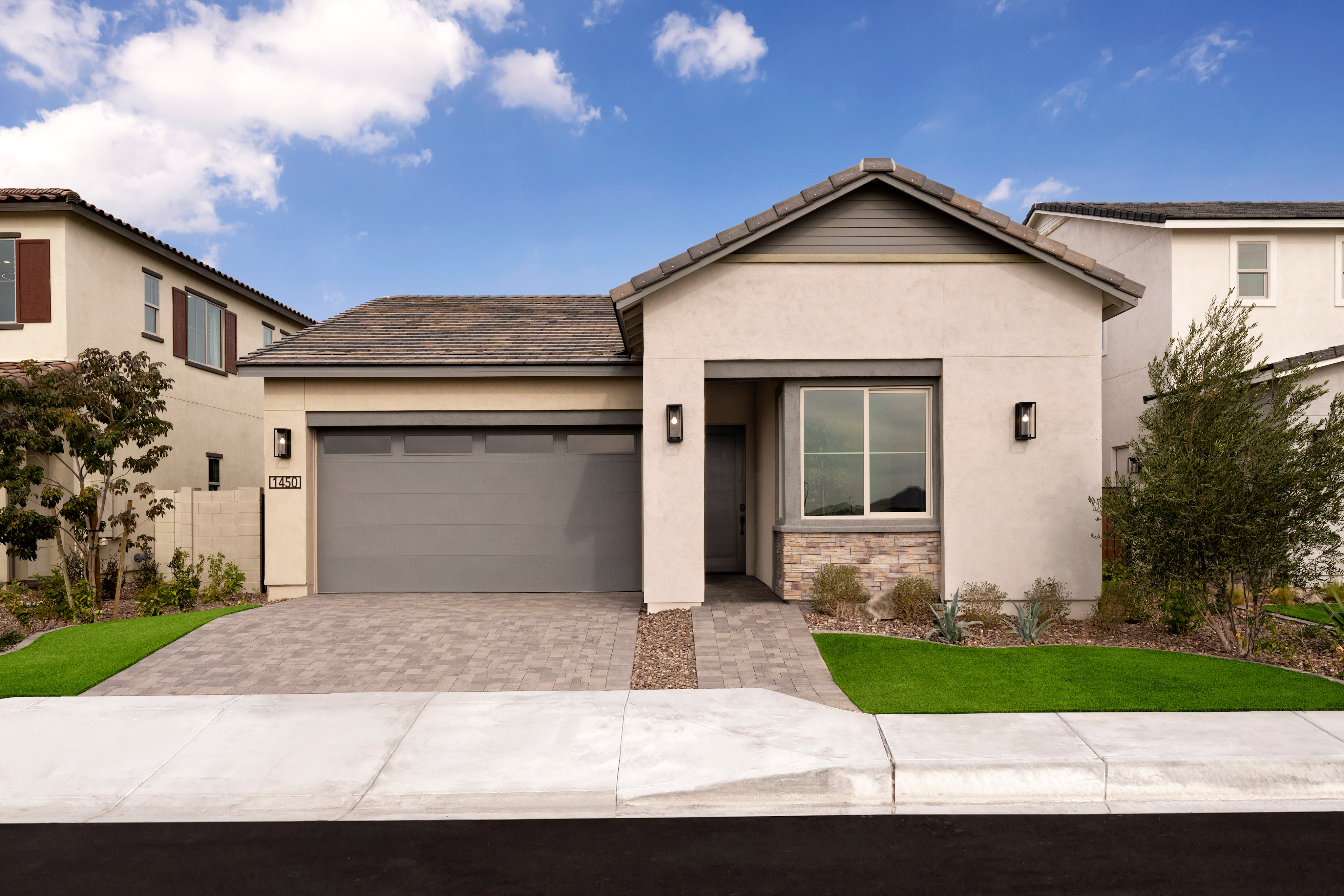 A modern, single-story house with a garage, surrounded by a well-manicured lawn and landscaping, set against a blue sky with fluffy white clouds.