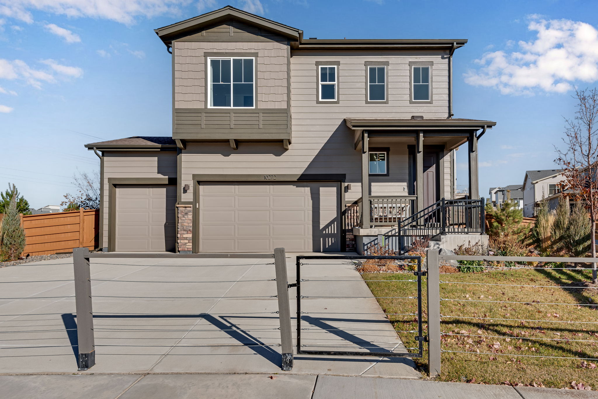 A two-story residential house with a gray exterior, a covered porch, and a fenced yard in the foreground, set against a backdrop of a clear sky with some clouds.