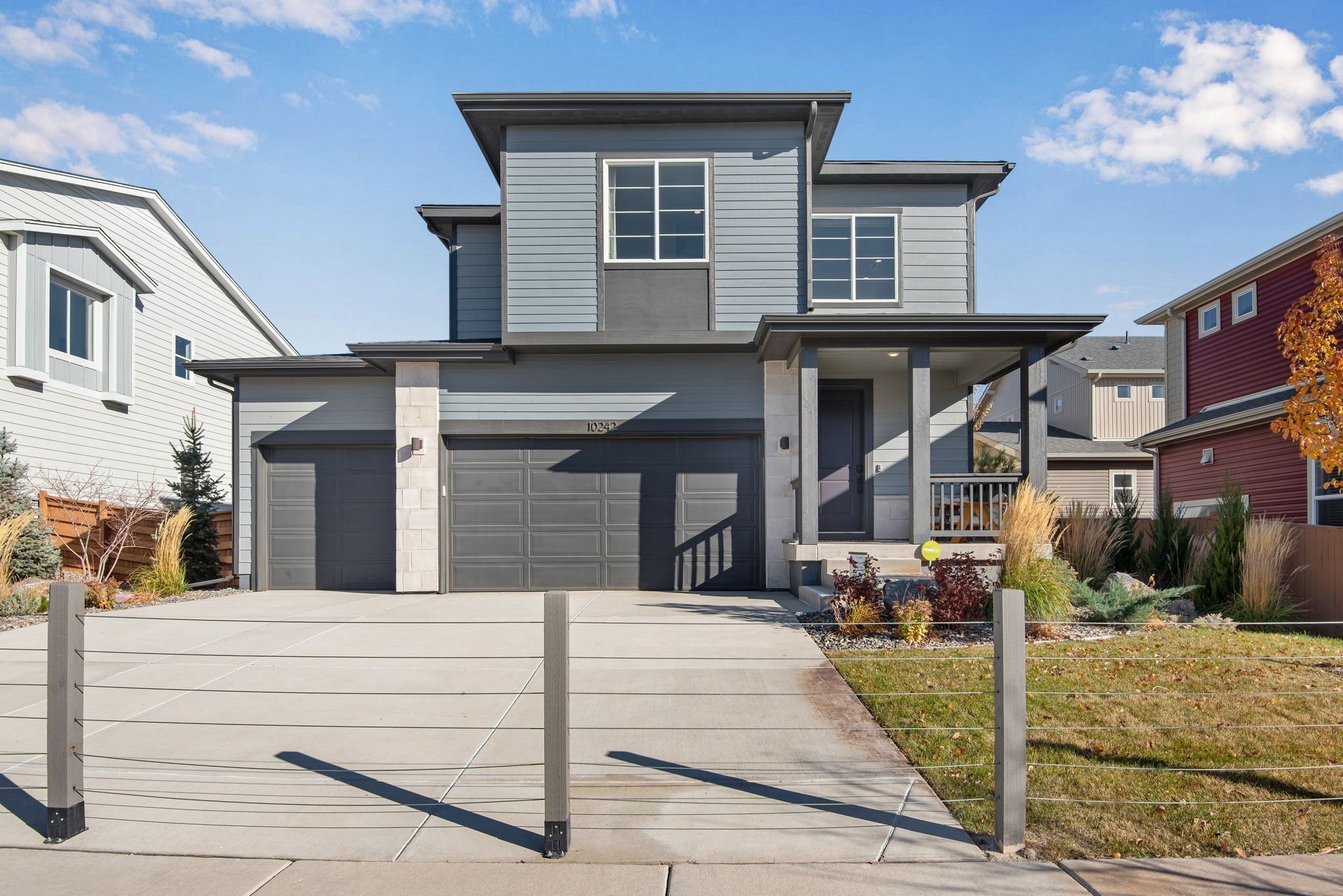 A modern two-story house with a gray exterior, a garage, and a well-maintained yard with a paved walkway leading to the front entrance, set against a backdrop of a clear blue sky with scattered clouds.