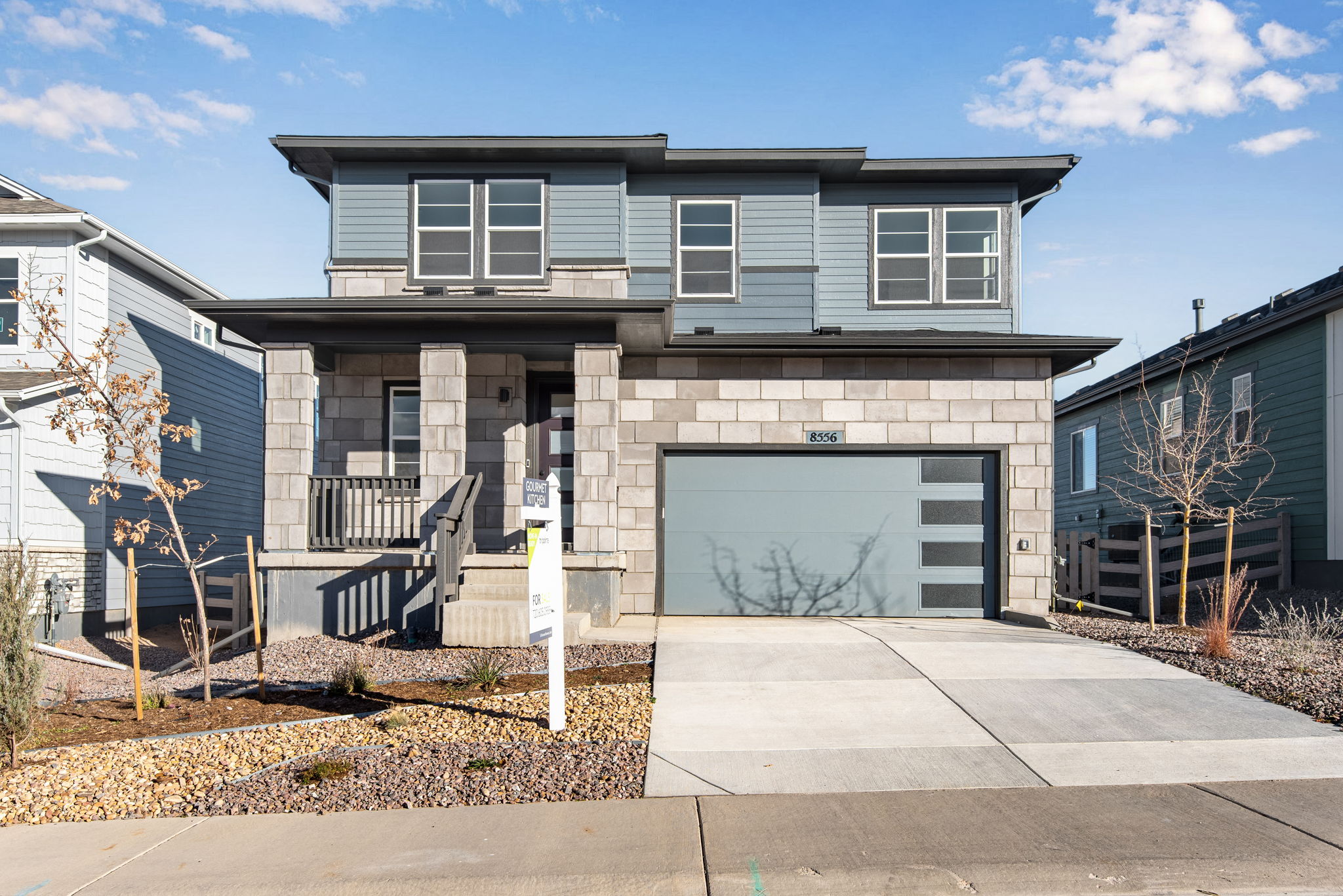 A modern two-story residential building with a gray exterior, large windows, and a paved driveway leading to the front entrance, set against a backdrop of a clear sky with scattered clouds.