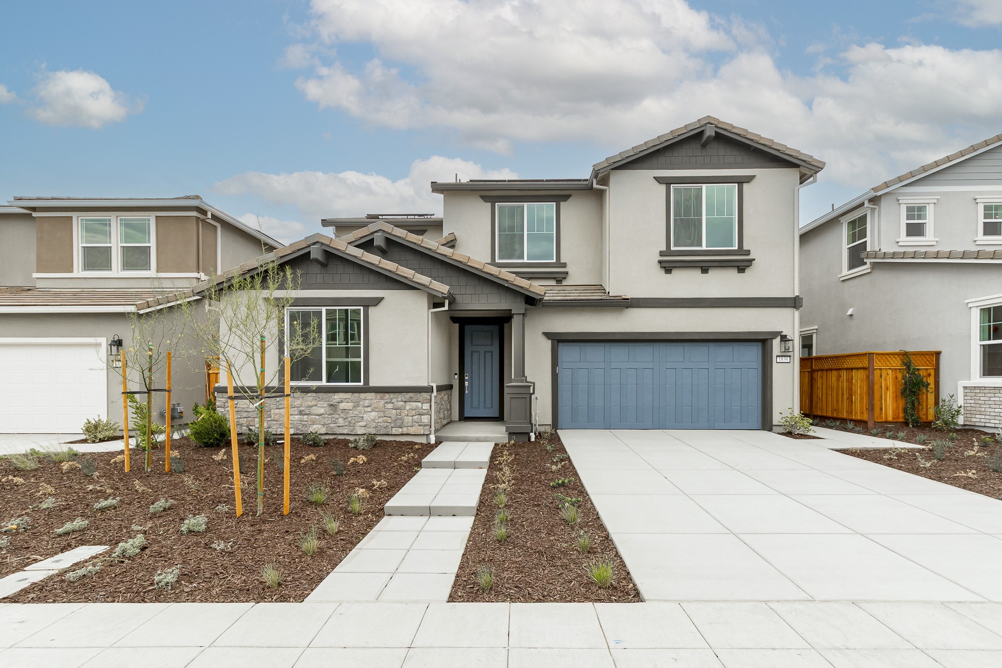 A modern two-story residential house with a garage, surrounded by a paved driveway and landscaped yard, set against a backdrop of cloudy blue sky.