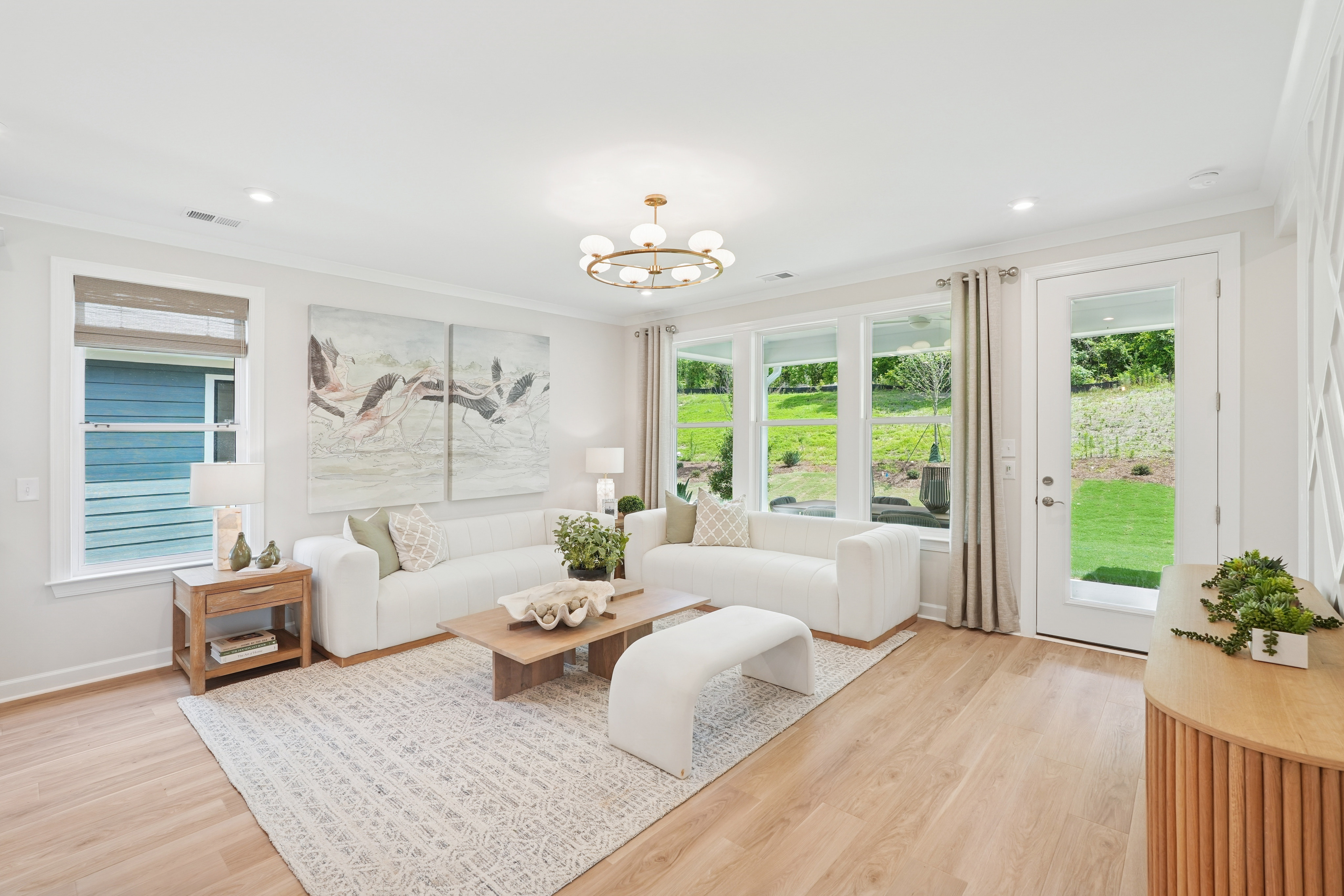 A bright and airy living room with white furniture, a large window overlooking a lush green backyard, and a decorative wall art piece.