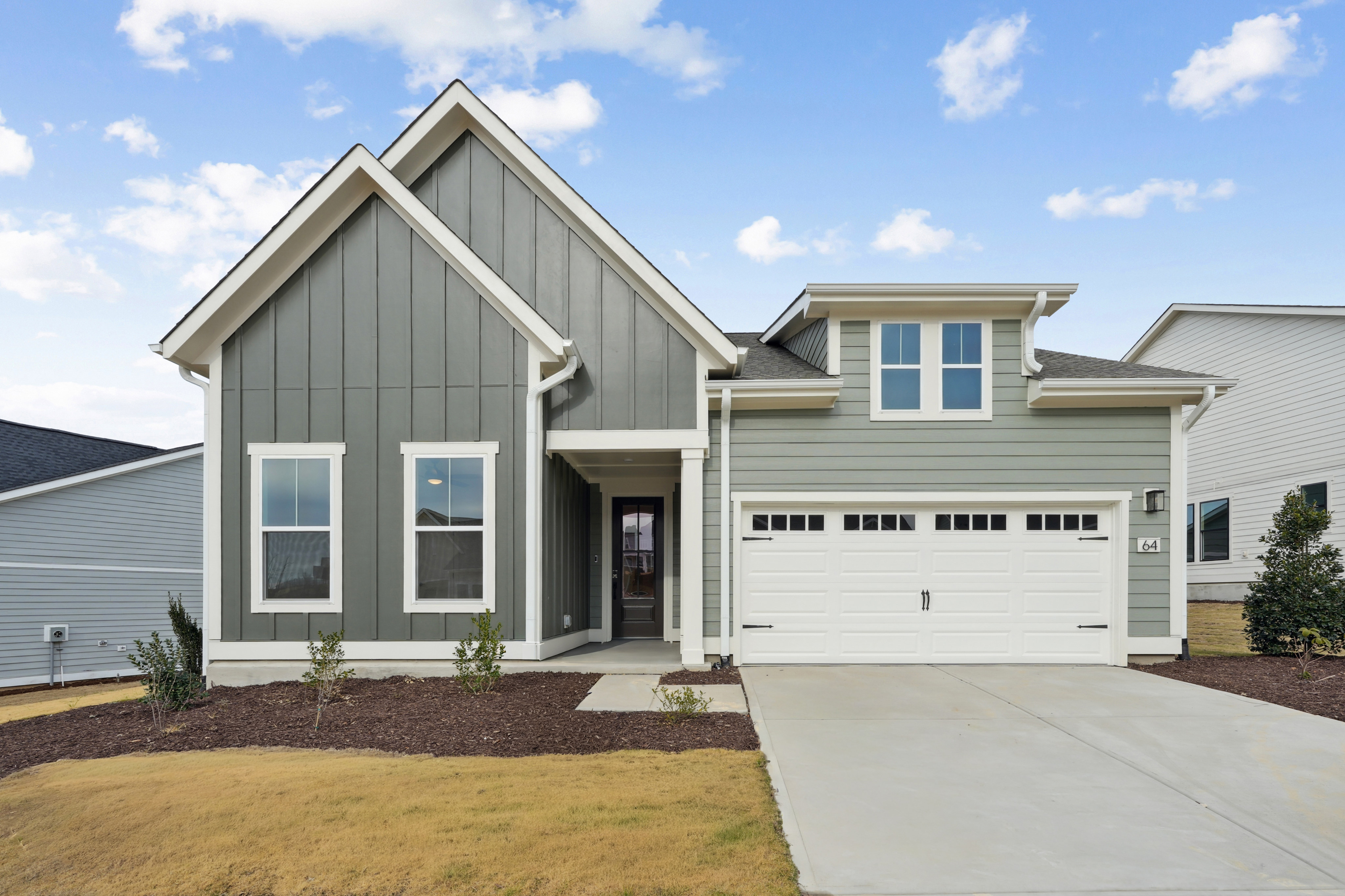 A modern two-story house with a gray exterior, a gabled roof, and a garage door stands in a grassy yard against a backdrop of a blue sky with fluffy white clouds.