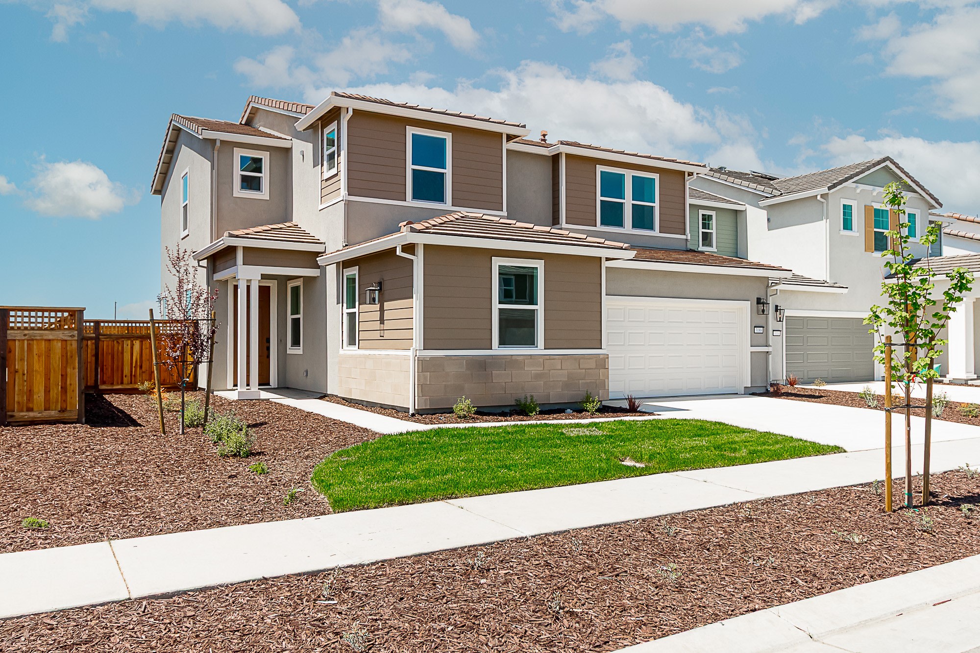 A two-story residential house with a well-manicured lawn, surrounded by a paved driveway and a wooden fence, set against a backdrop of a blue sky with fluffy white clouds.