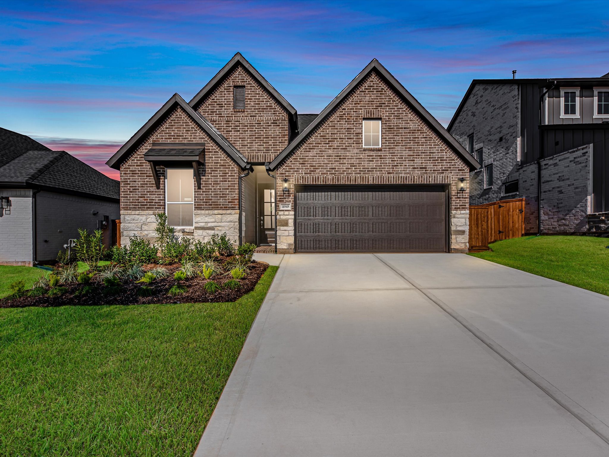 A modern two-story brick house with a garage and a well-manicured lawn in the foreground, set against a vibrant sky with a colorful sunset in the background.