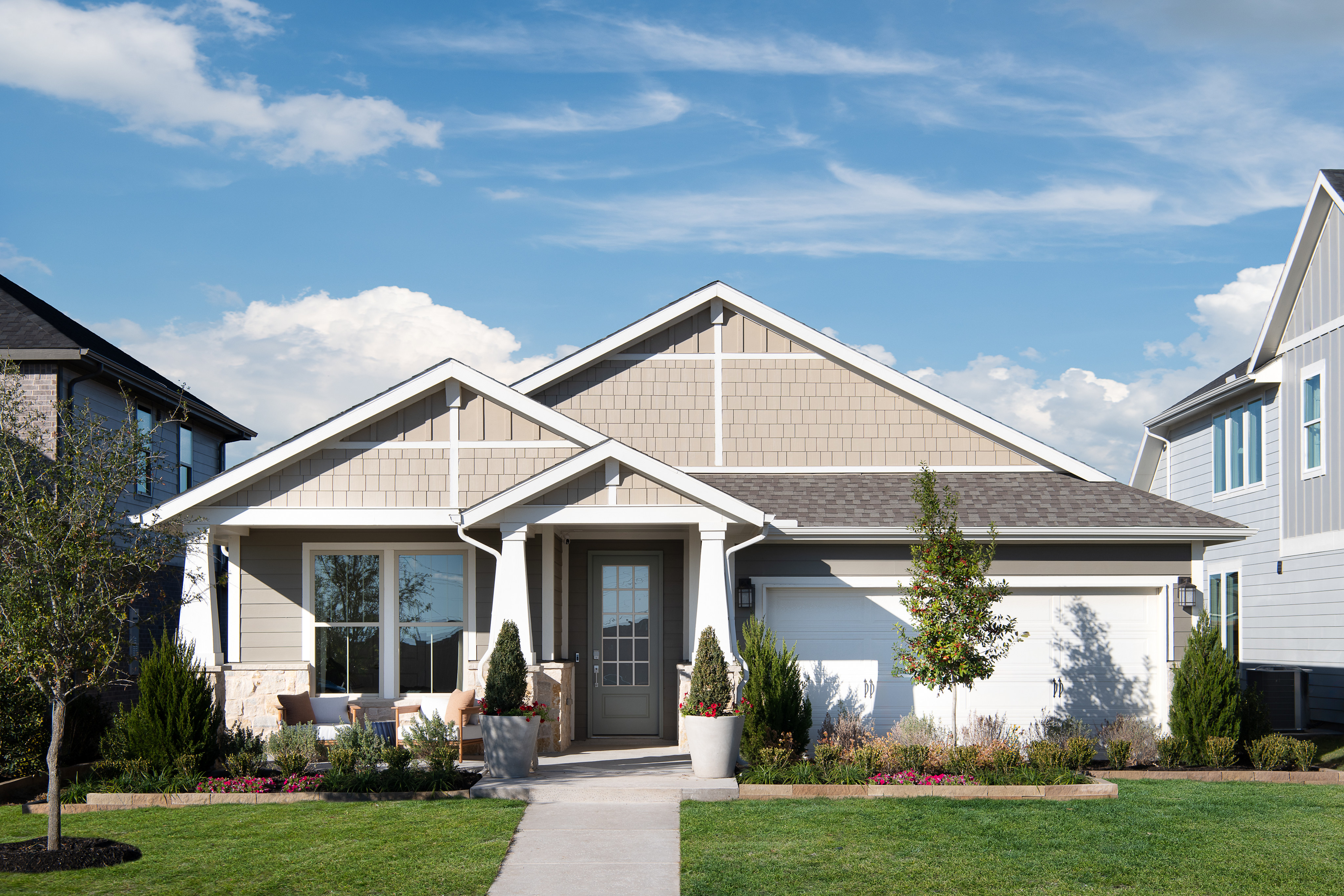 A well-manicured lawn leads to a modern, two-story house with a gabled roof, siding, and large windows, set against a backdrop of a blue sky with wispy clouds.