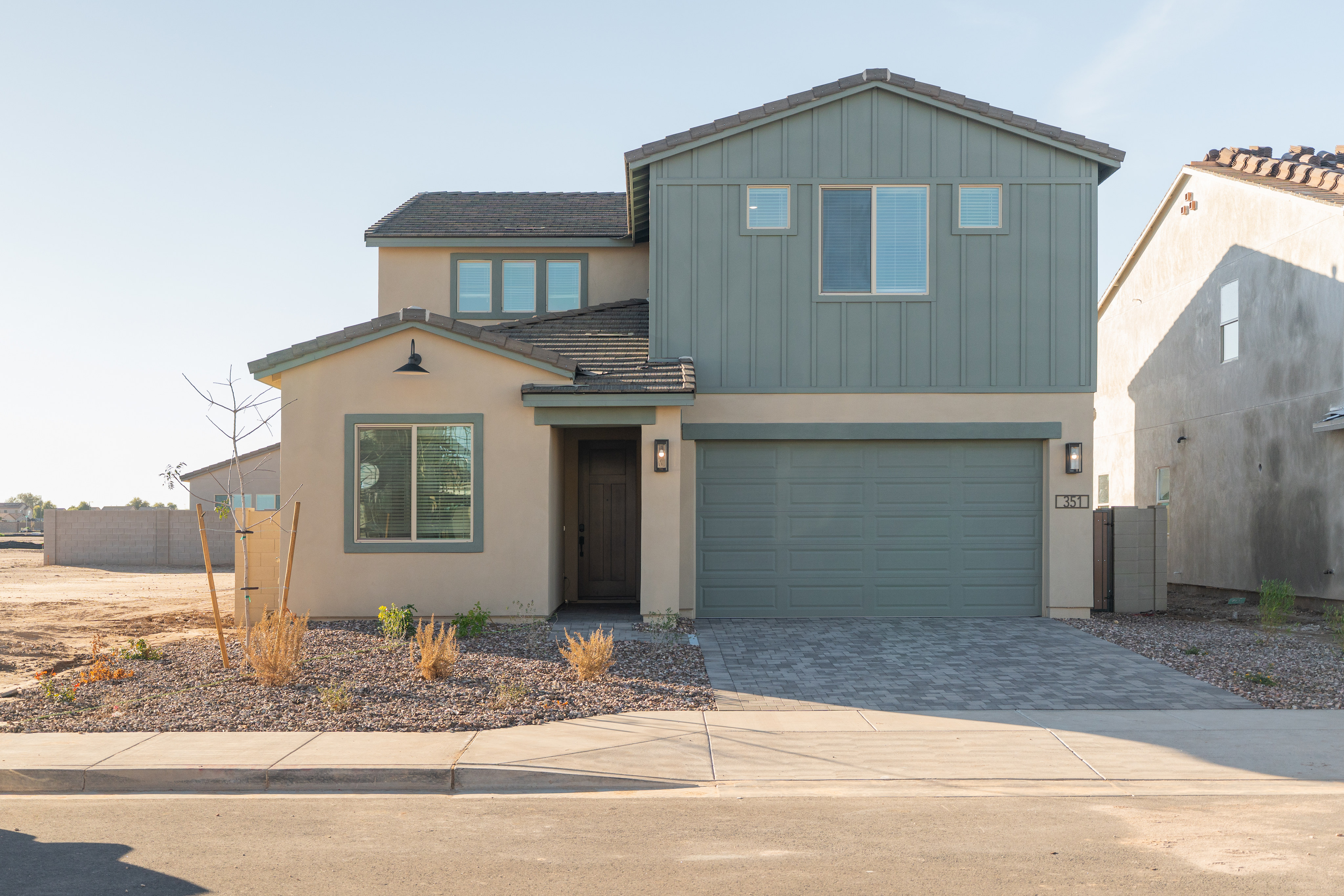 A modern two-story house with a garage, surrounded by a paved driveway and landscaping, stands against a clear blue sky.