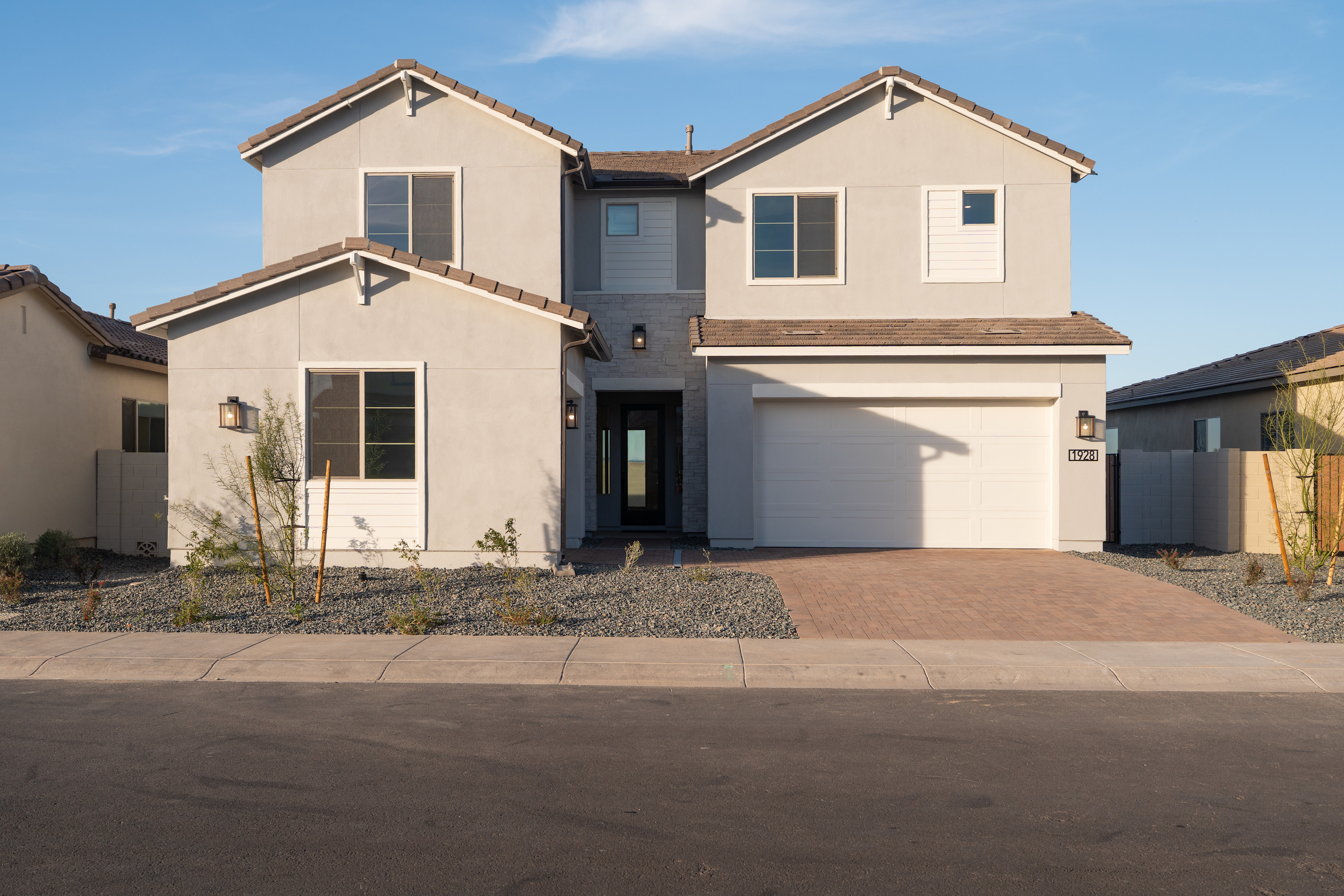 A two-story beige house with a garage and a paved driveway, set against a clear blue sky with some clouds.