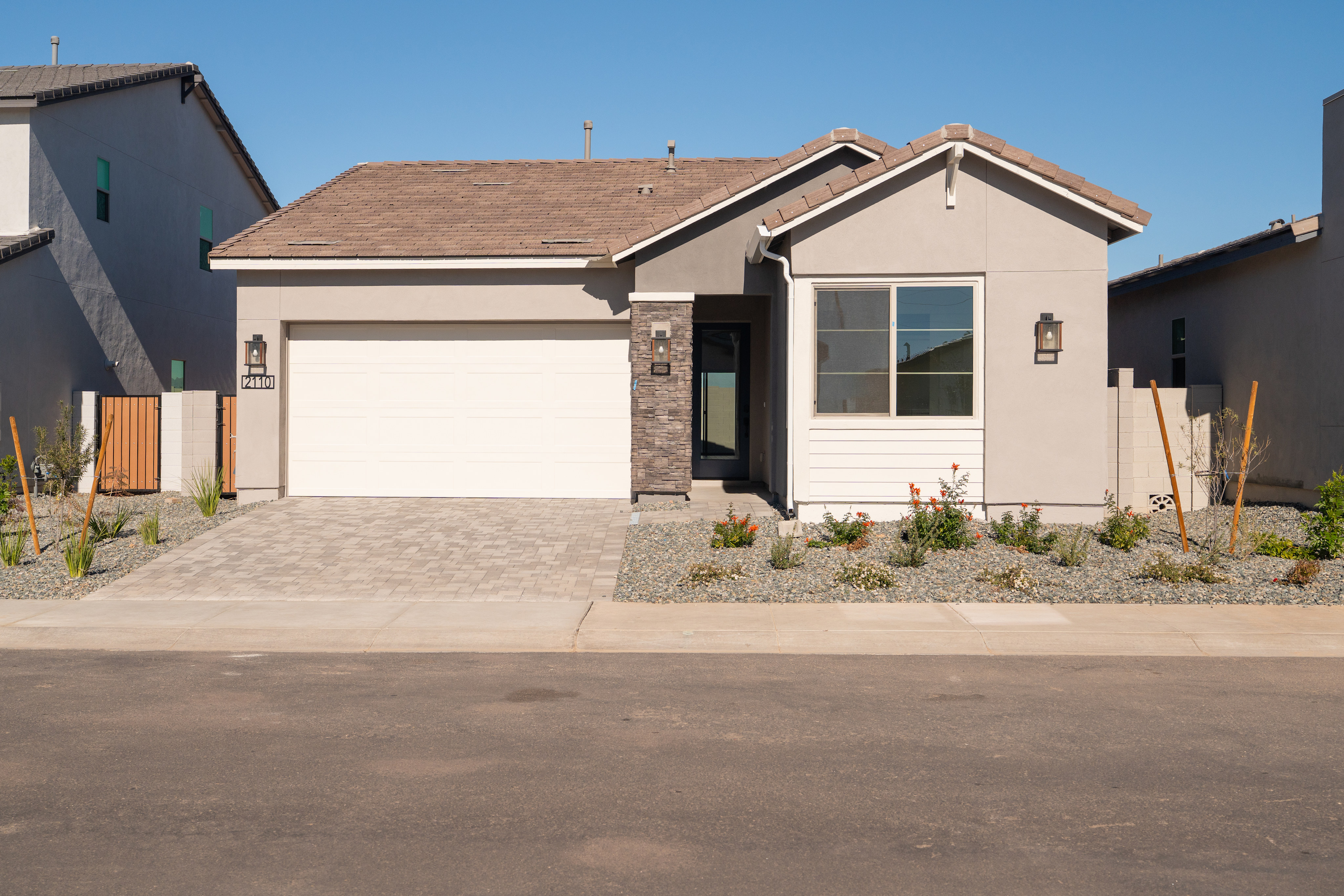 A single-story residential house with a garage, surrounded by a paved driveway and landscaped yard under a clear blue sky.