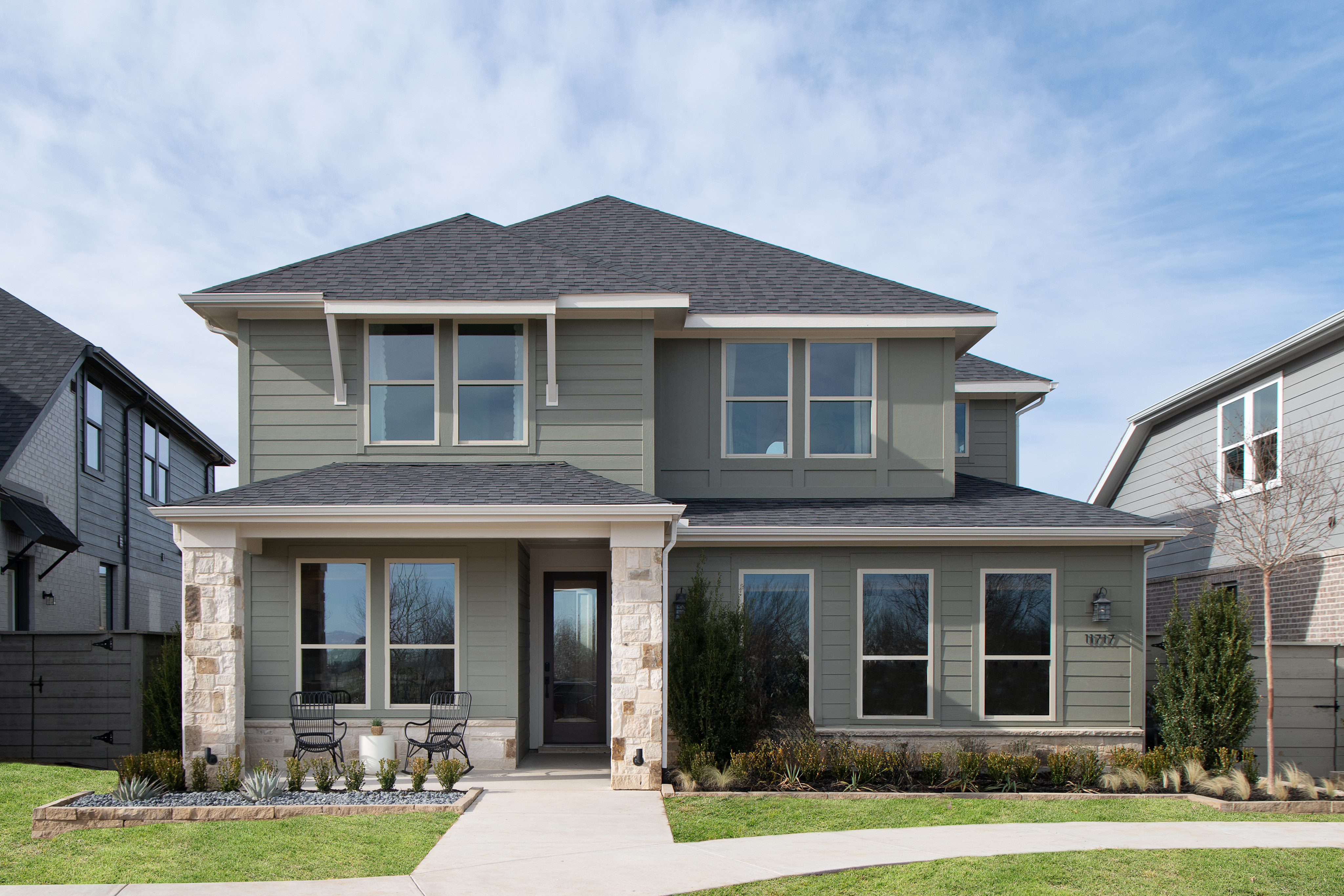 A two-story residential house with a gray exterior, a slanted roof, and large windows, surrounded by a well-maintained lawn and landscaping in the foreground.