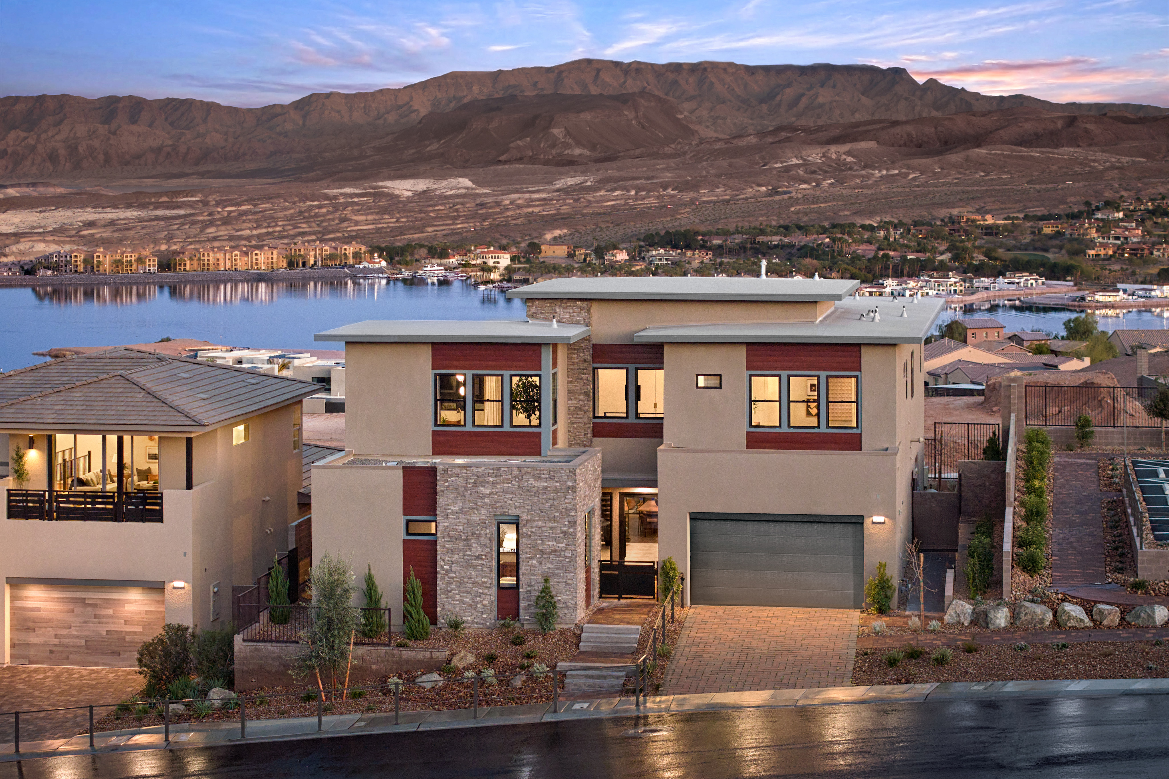 The image depicts a residential neighborhood nestled against a mountainous landscape, with a body of water in the foreground reflecting the warm hues of the sunset.
