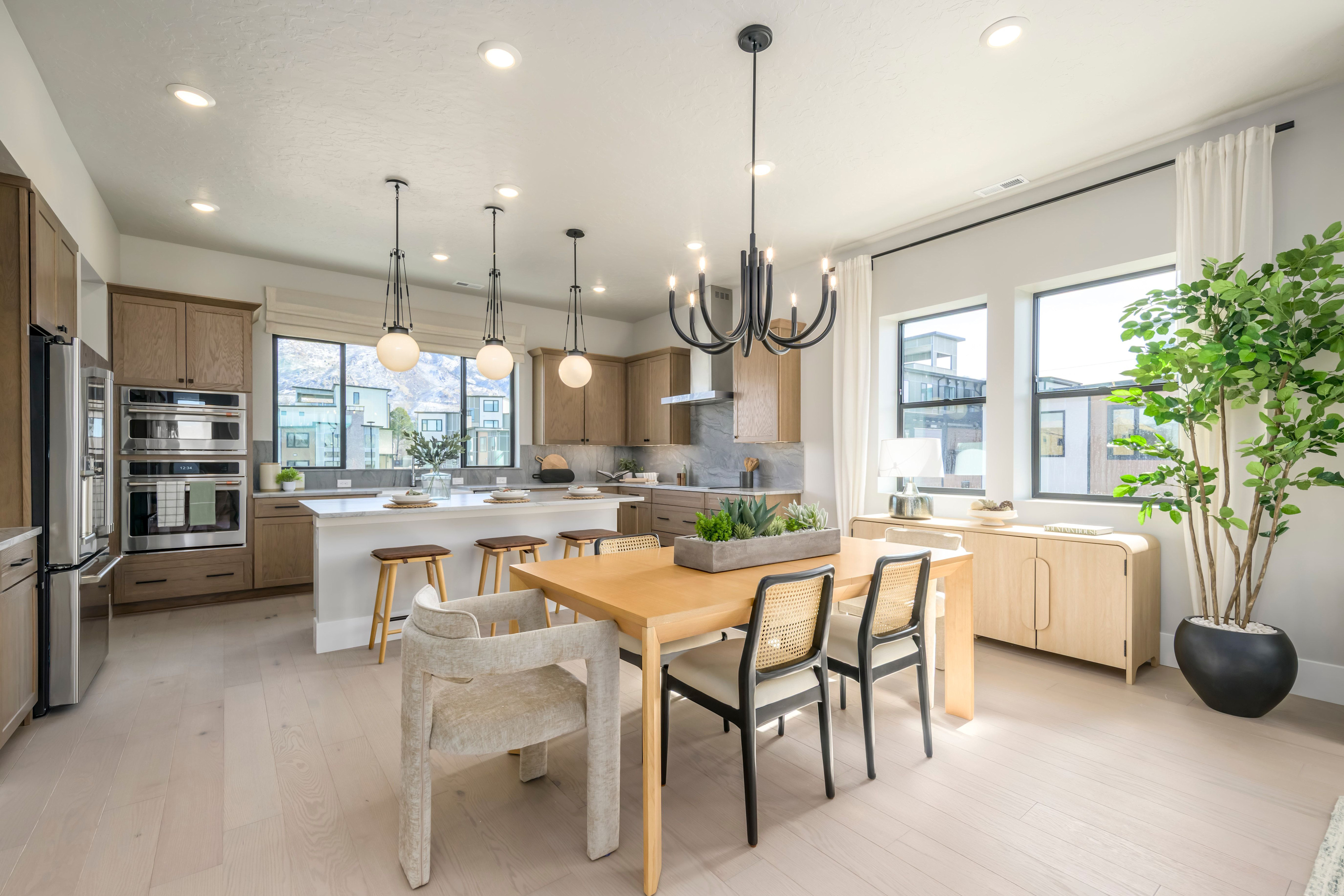 A modern and spacious kitchen with a dining area, featuring light-colored cabinets, a large island, and a variety of pendant lights and potted plants.
