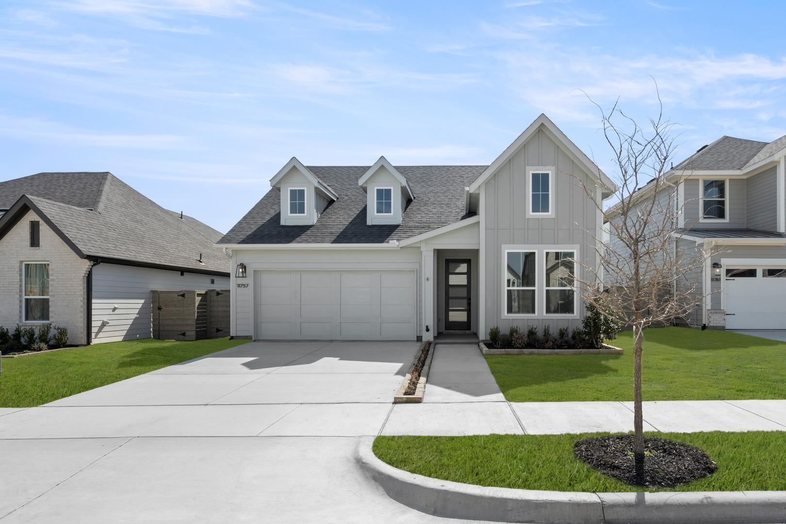 A two-story residential house with a gray roof, white siding, and a well-manicured lawn in the foreground, set against a backdrop of other similar houses in a suburban neighborhood.