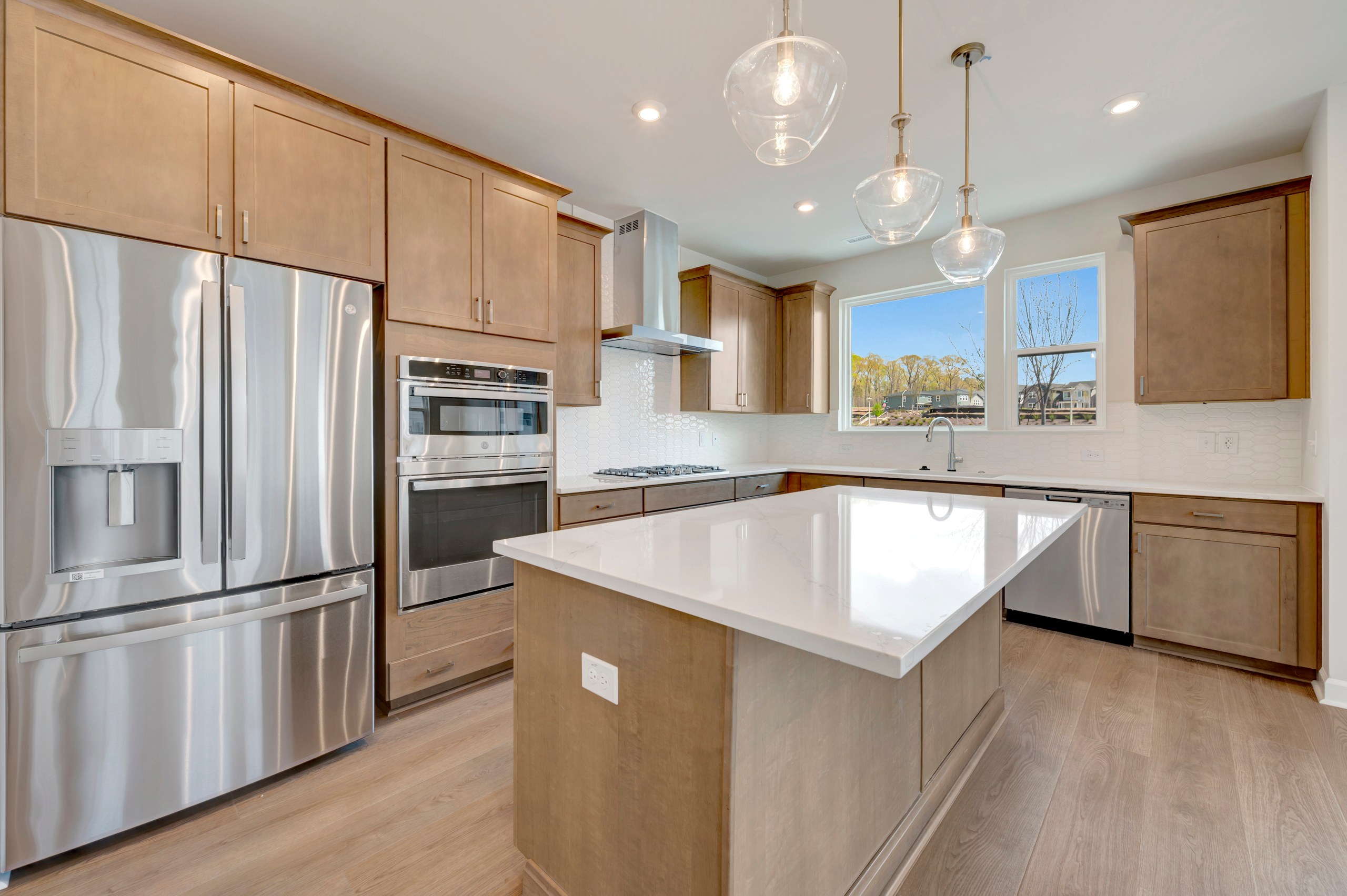 A modern, well-lit kitchen with light-colored wood cabinets, stainless steel appliances, and a central island with a white countertop.