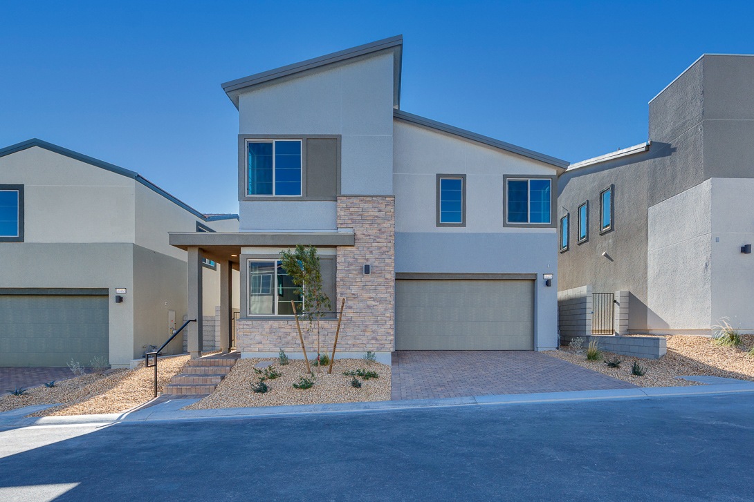 A modern, two-story residential building with a garage, surrounded by a paved driveway and landscaped yard against a clear blue sky.