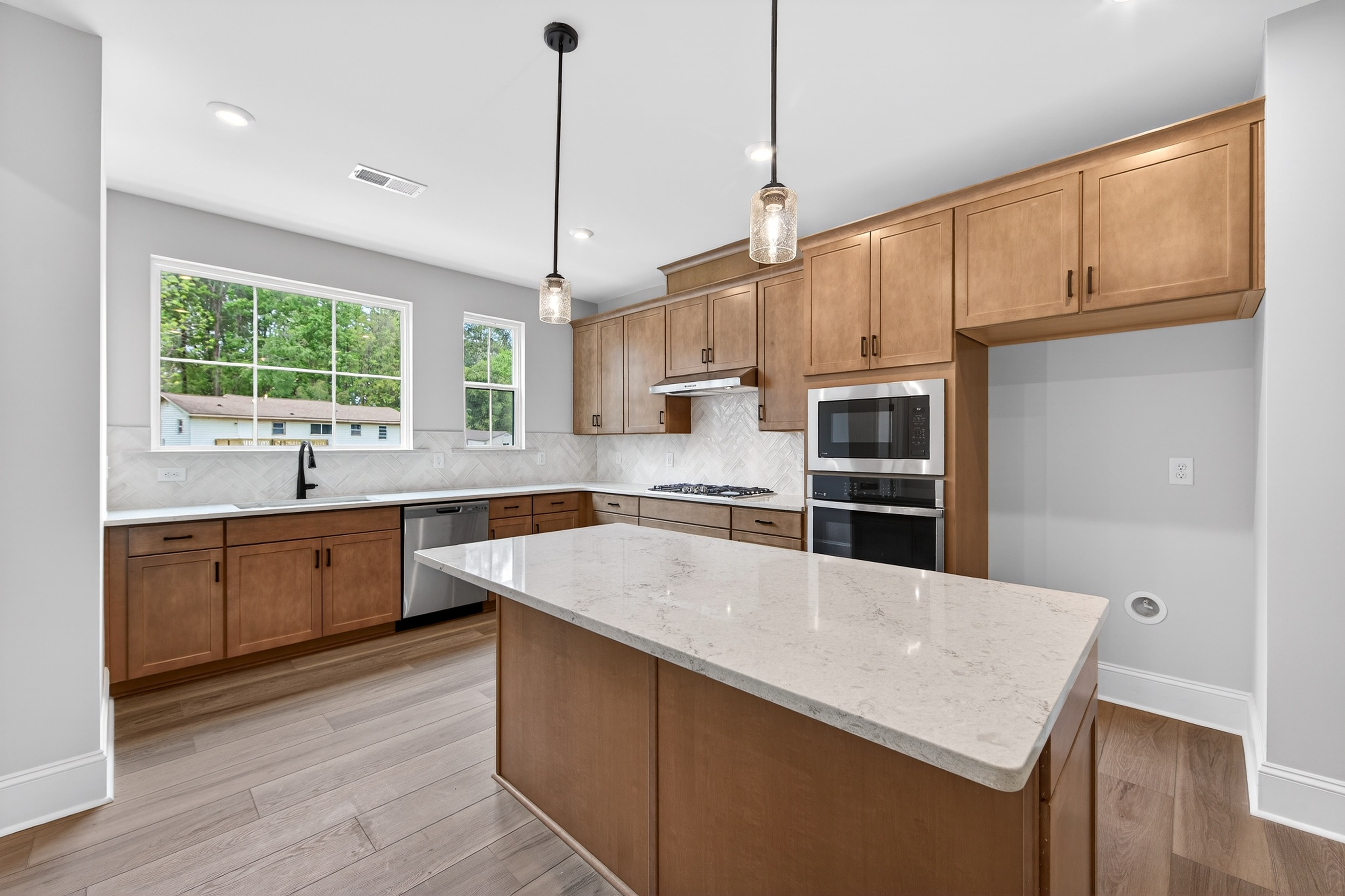 A modern, well-lit kitchen with wooden cabinets, white countertops, and large windows overlooking a lush outdoor scene.