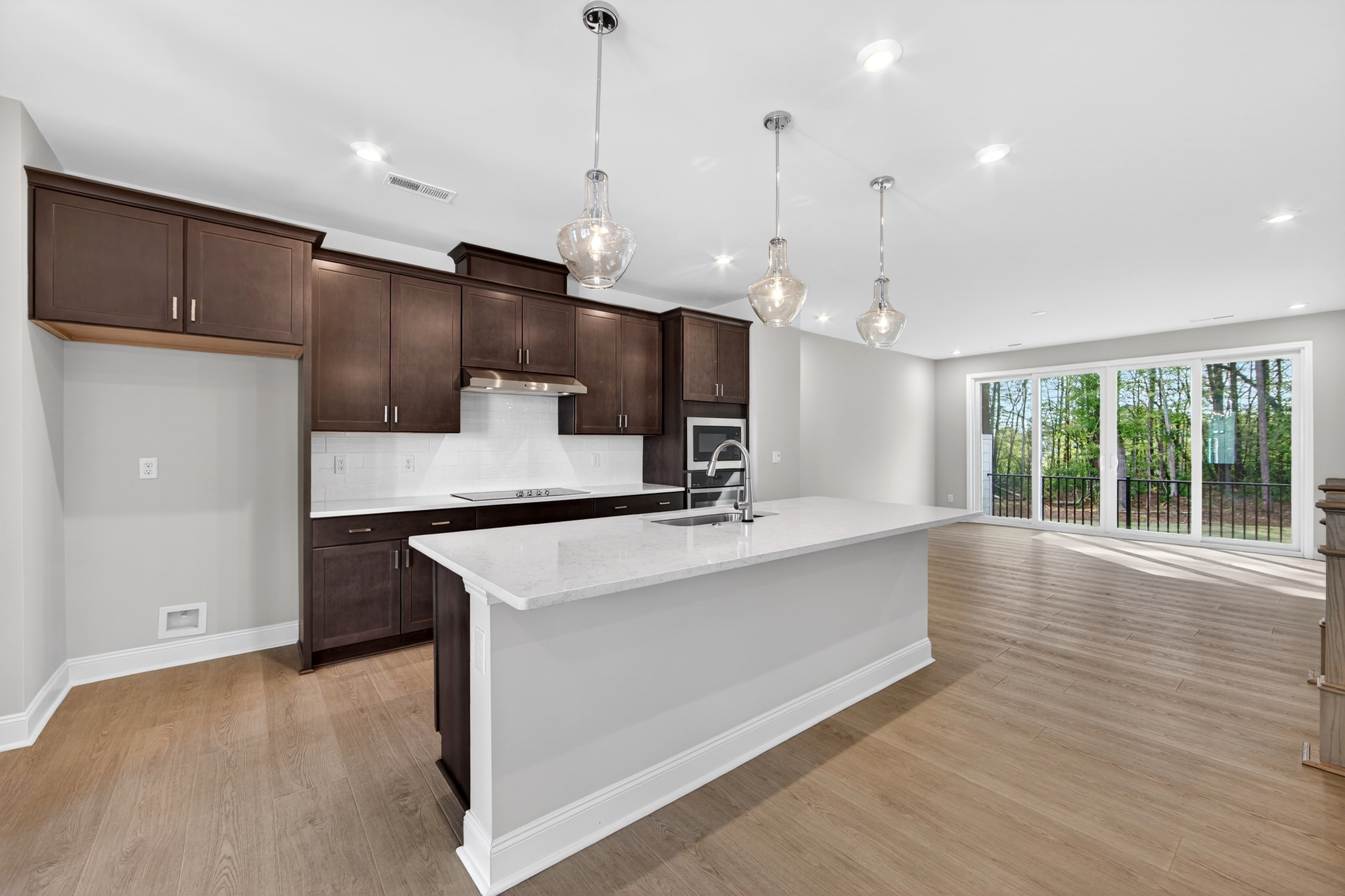 A modern, open-concept kitchen with dark wood cabinets, white countertops, and pendant lighting, leading into a spacious living area with hardwood floors and large windows overlooking a wooded outdoor scene.