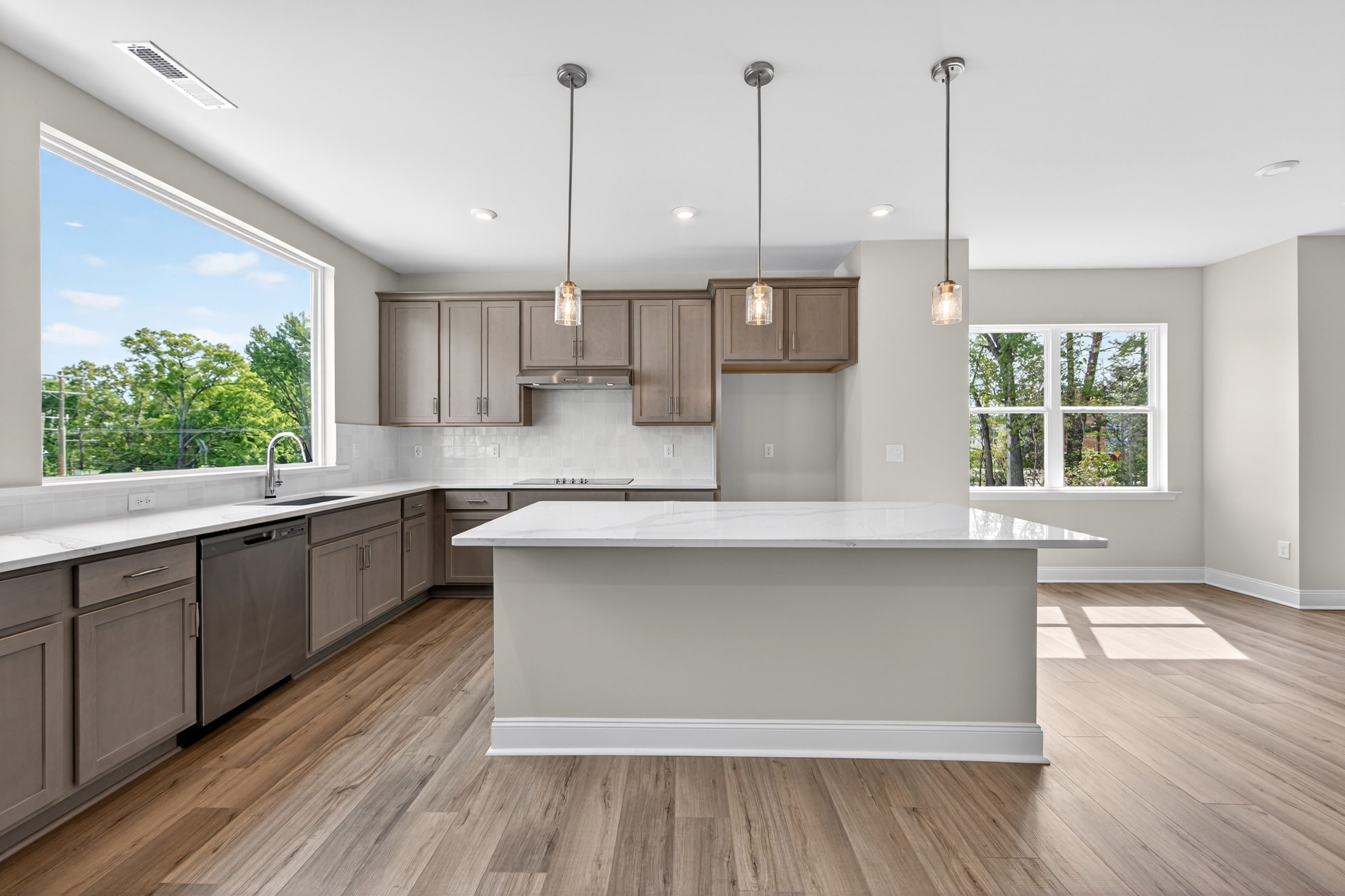 A modern, open-concept kitchen with gray cabinets, a white island, and large windows overlooking a wooded outdoor area.