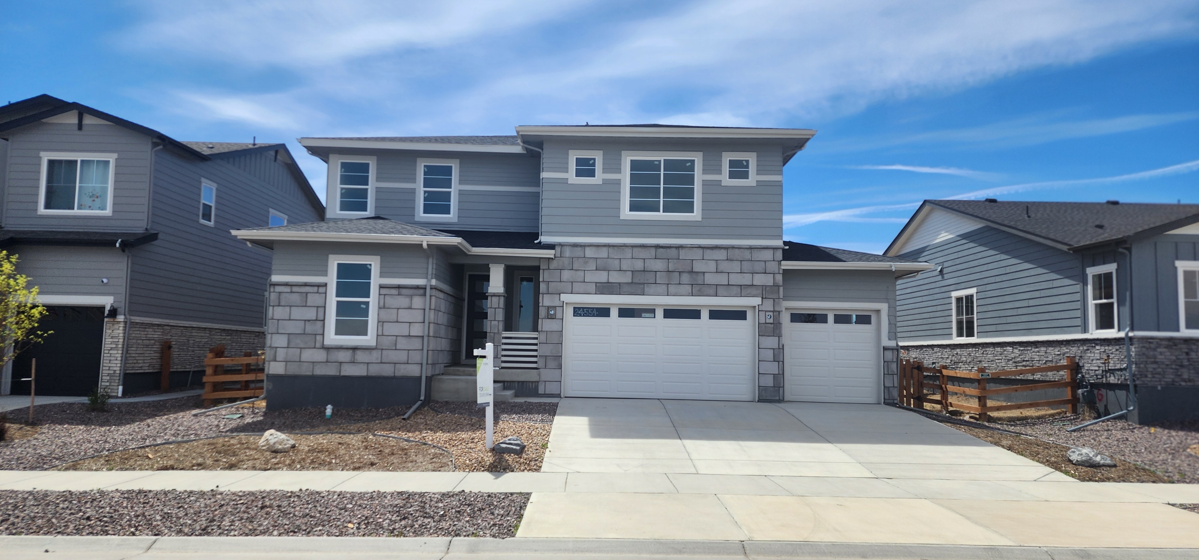 A modern two-story residential house with a gray exterior, a garage, and a paved driveway, set against a blue sky with scattered clouds.