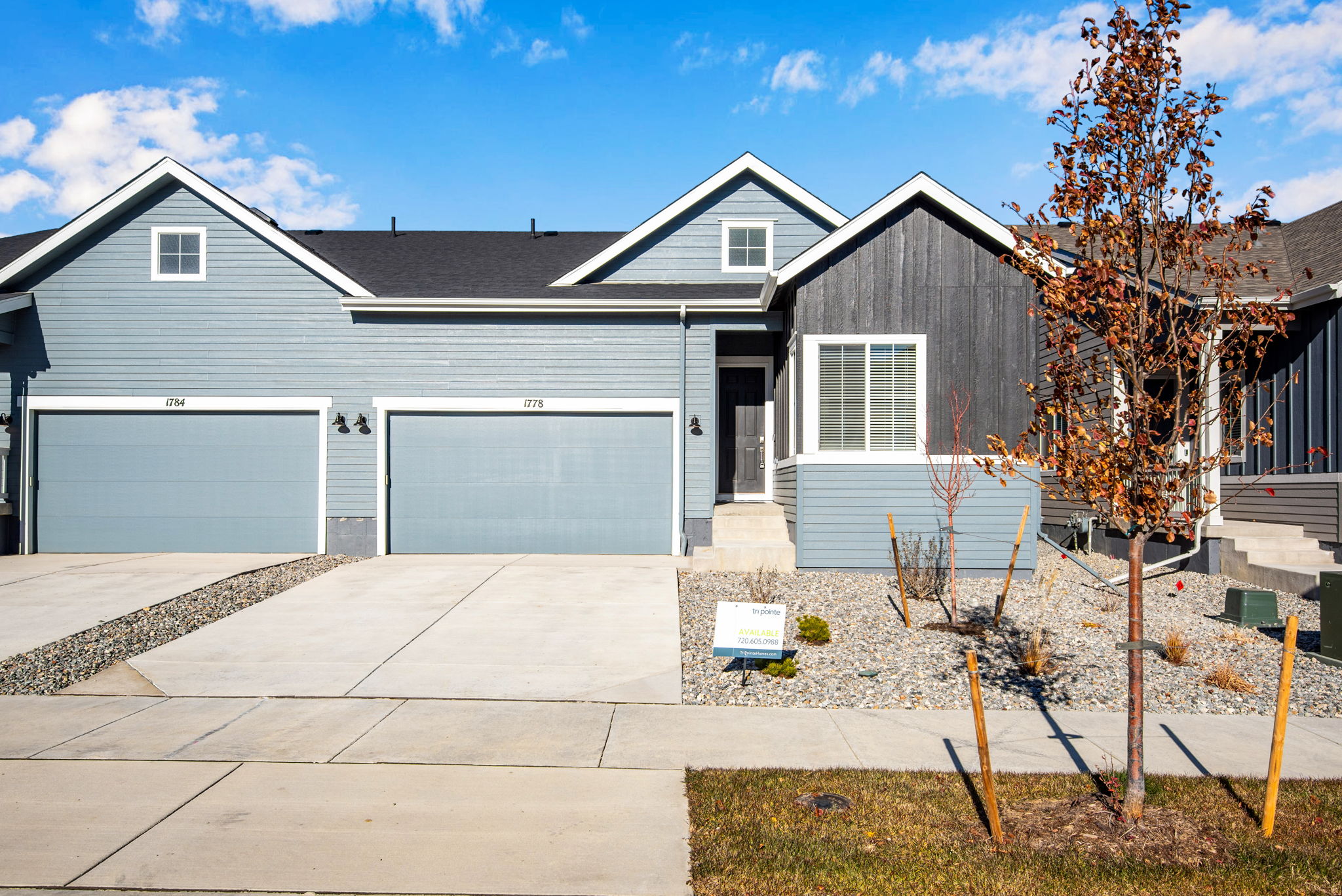 A modern two-story house with a gray exterior, a slanted roof, and a paved driveway leading to a garage. The house is surrounded by a grassy lawn and a tree with autumn-colored leaves in the background.