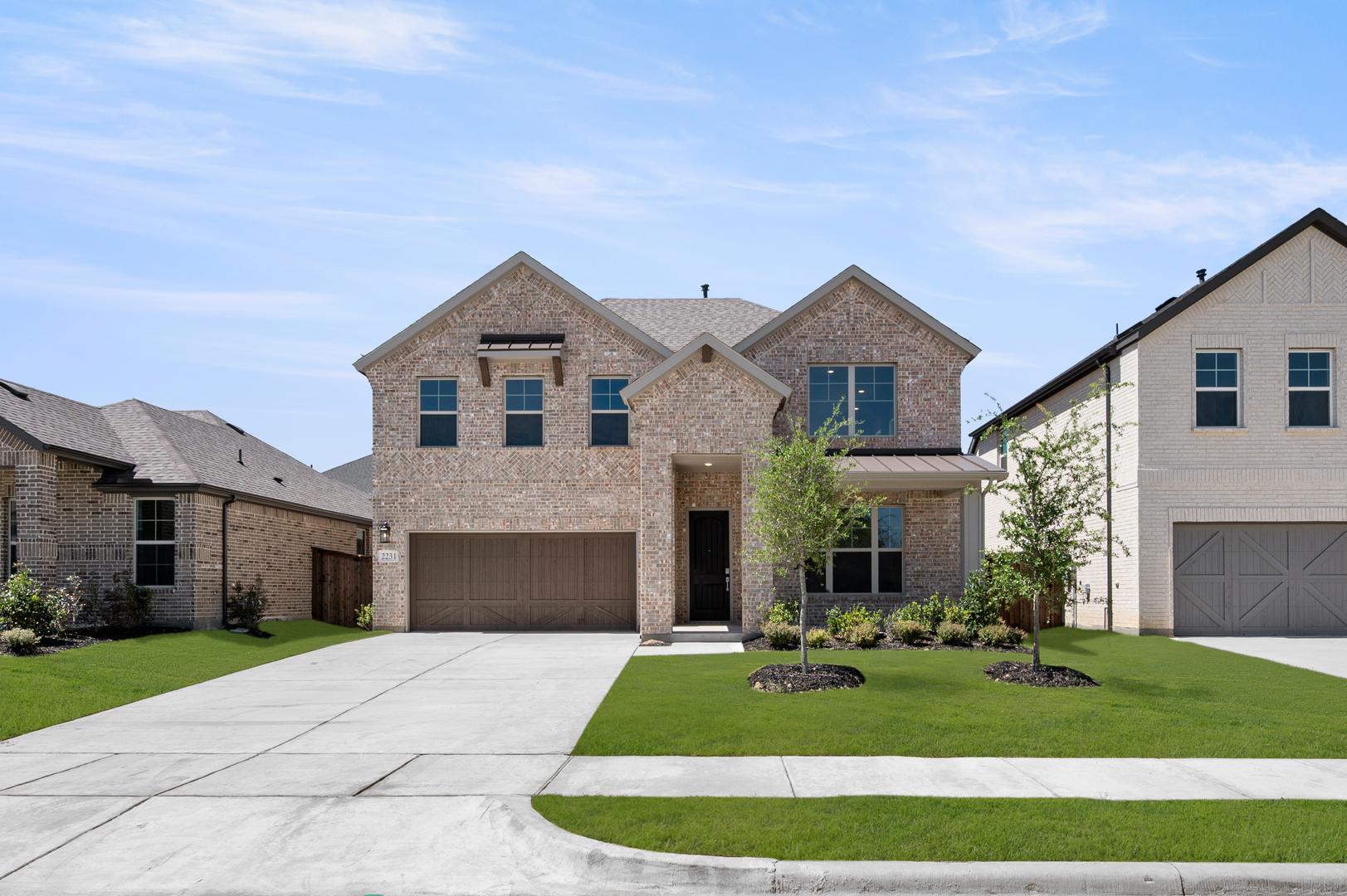 A two-story brick house with a gabled roof, surrounded by a well-manicured lawn and a paved driveway, set against a clear blue sky.