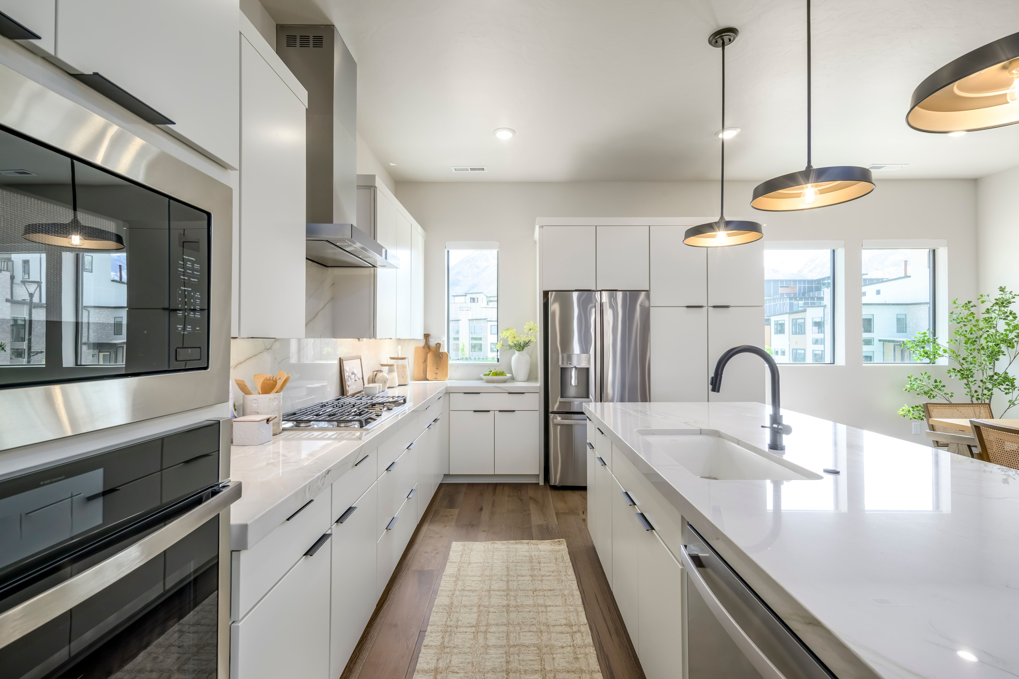 A modern white kitchen with a large island in the foreground and windows overlooking a residential area in the background.