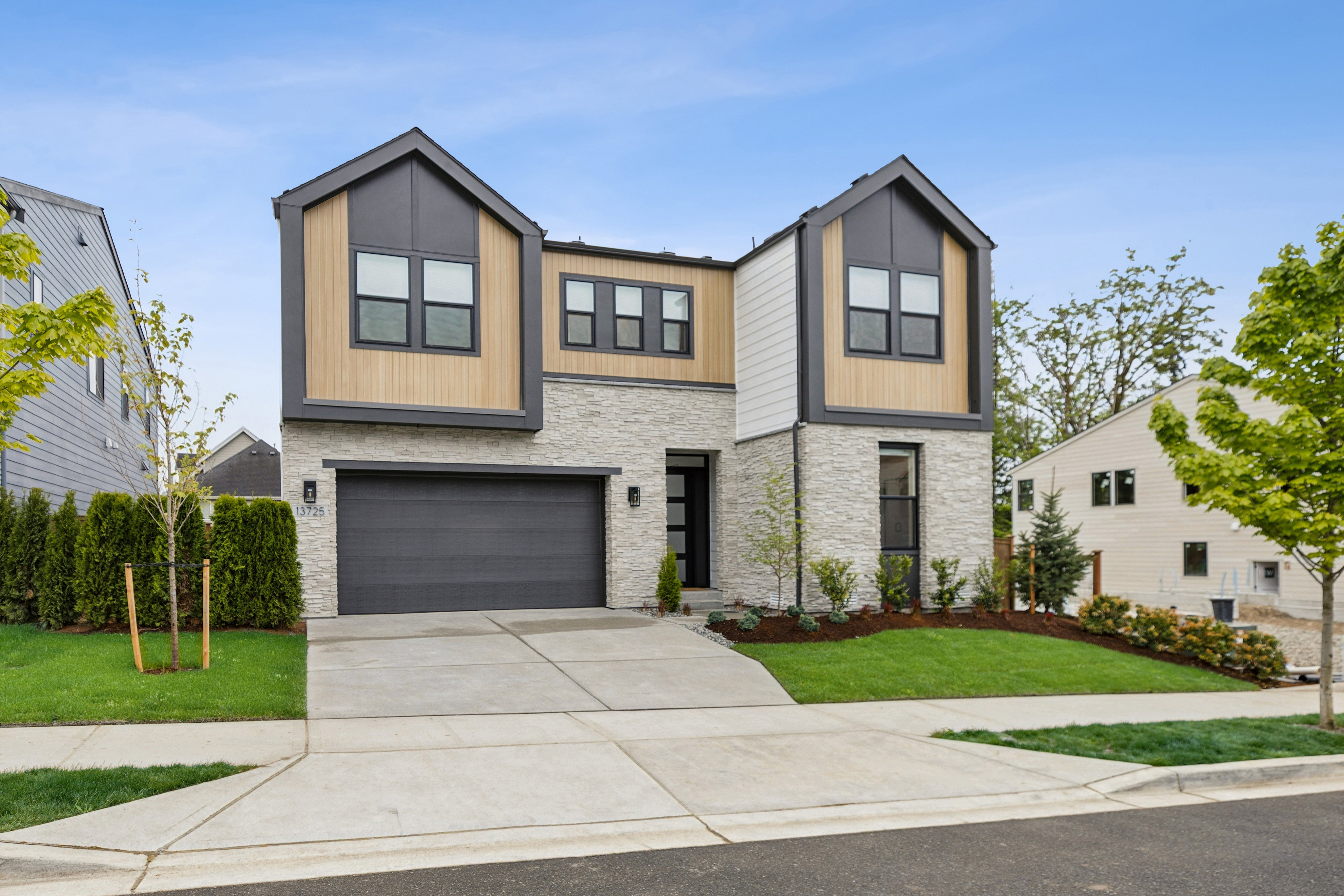 A modern two-story house with a gray garage door and stone accents sits on a manicured lawn, with neighboring houses and trees visible in the background under a clear blue sky.