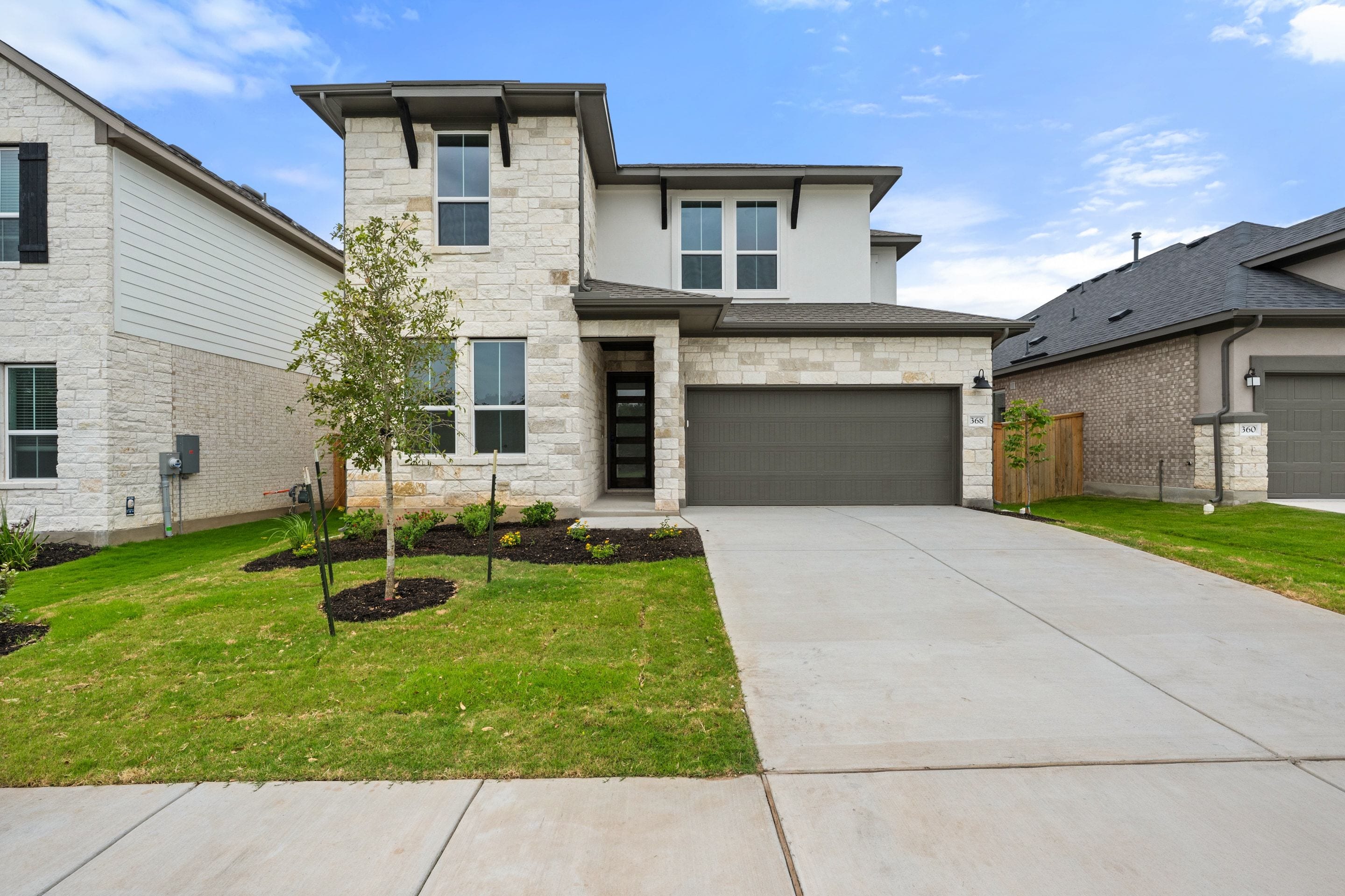 A modern two-story house with a light stone facade and a gray garage door sits on a manicured lawn, with neighboring houses and a cloudy blue sky in the background.