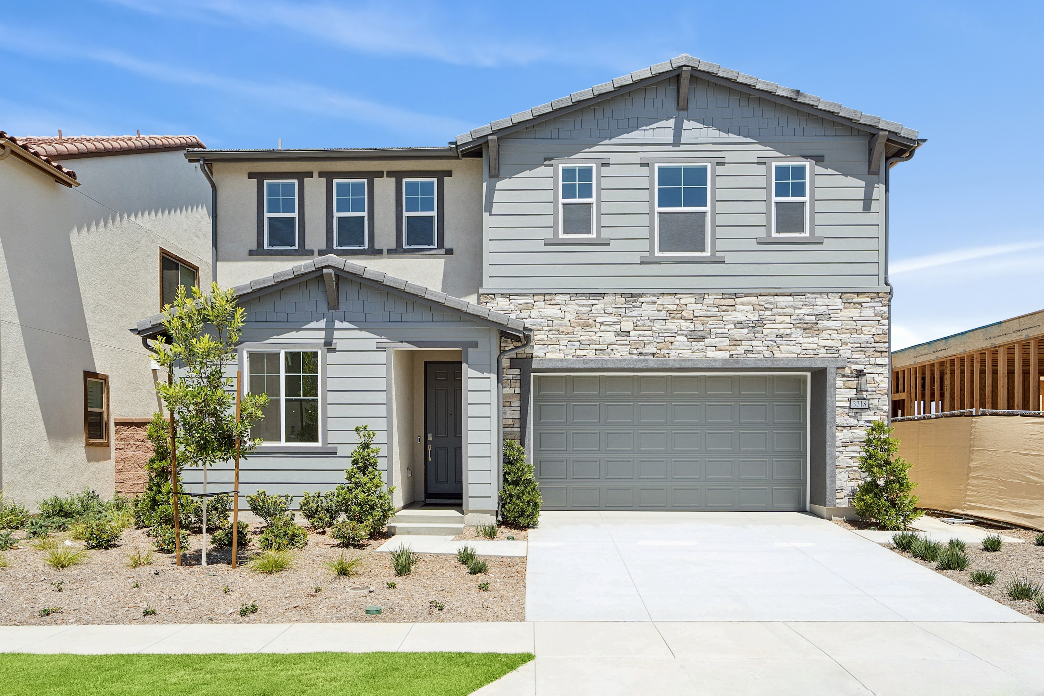 A modern two-story house with a stone accent facade and a gray garage door sits on a manicured lawn, with a clear blue sky and construction in the background.
