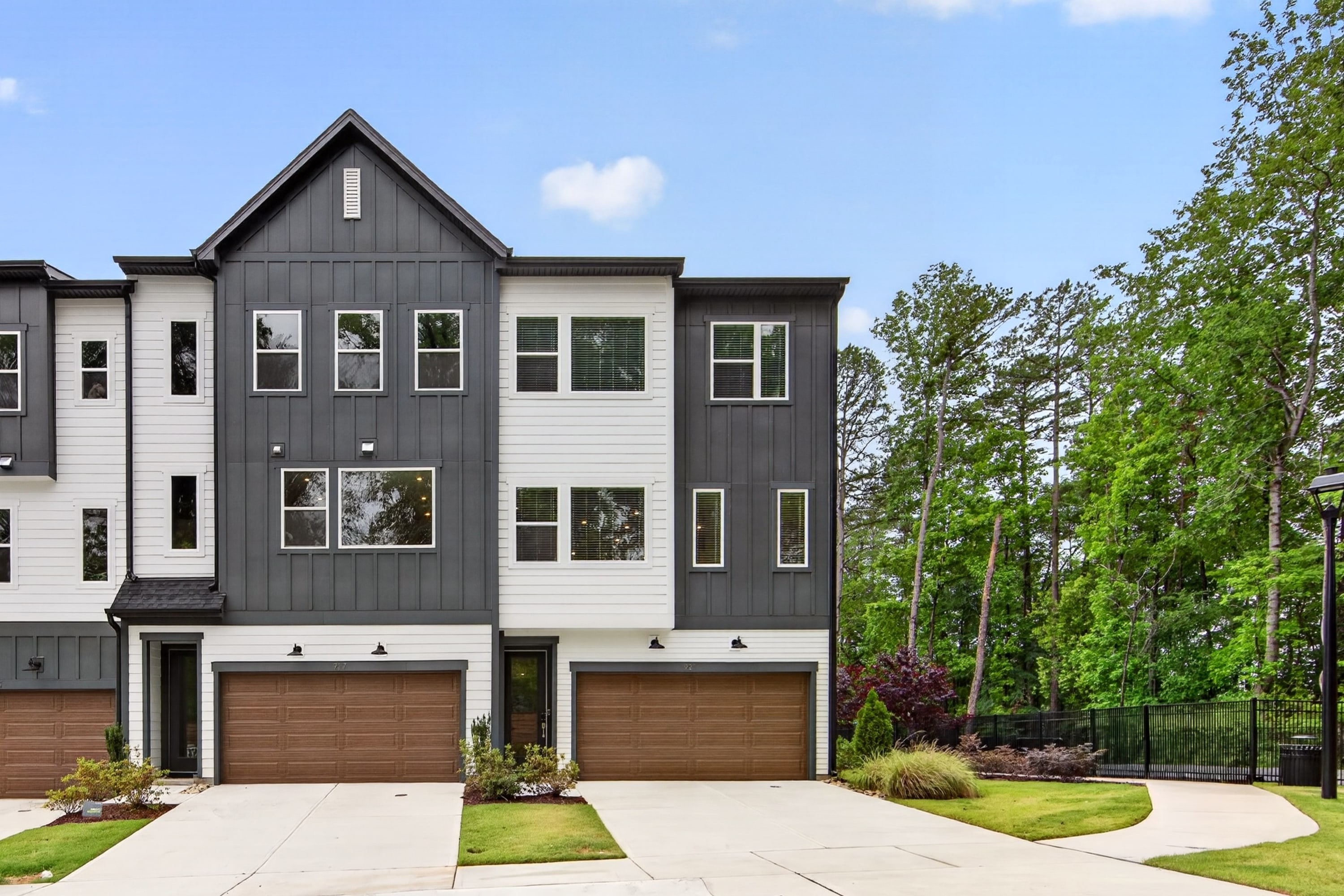 A modern townhouse with dark gray and white siding and brown garage doors stands in the foreground, backed by a lush green forest under a bright sky.